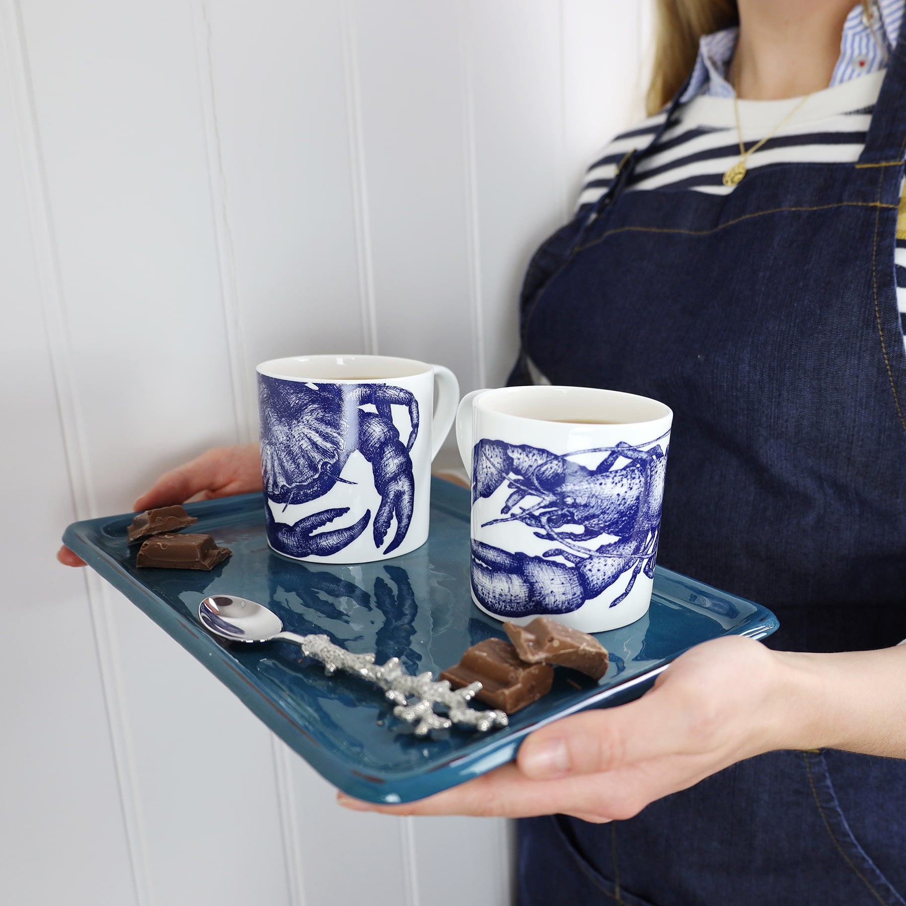 The photo shows Bone china white mug featuring hand drawn lobster design in classic Navy on a tray with a crab design mug,some chocolate and a pewter spoon