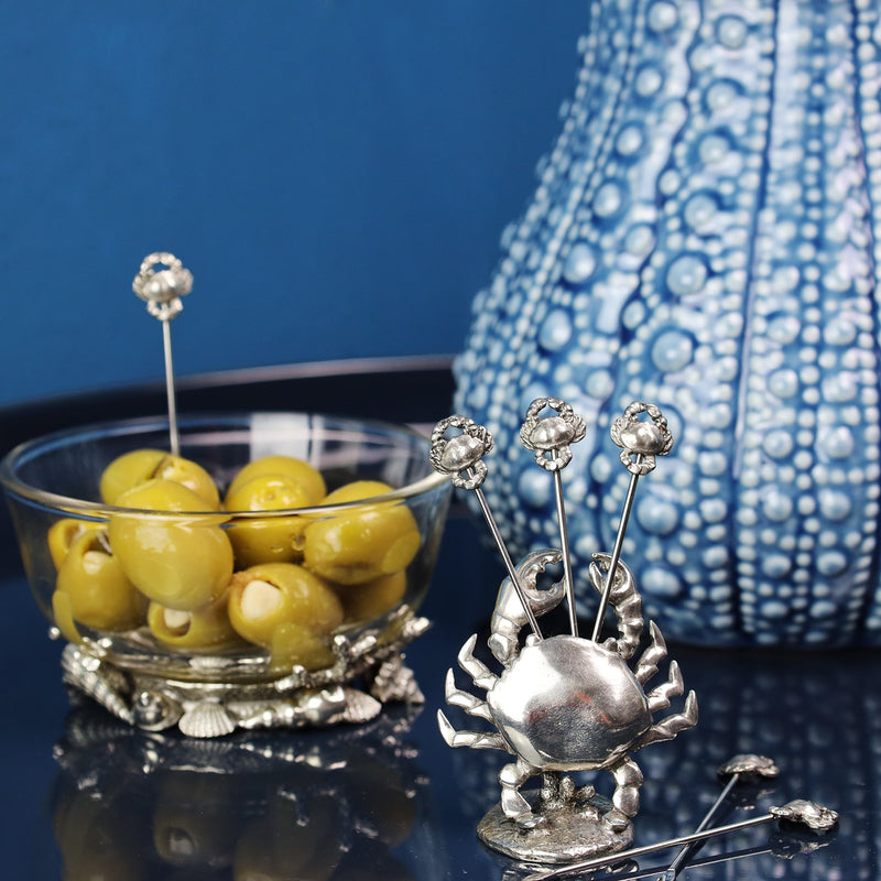 Pewter Crab shaped Pick Set with crabs at one end of each of the picks placed on a table in front of an urchin Jug and a condiment glass bowl containing olives