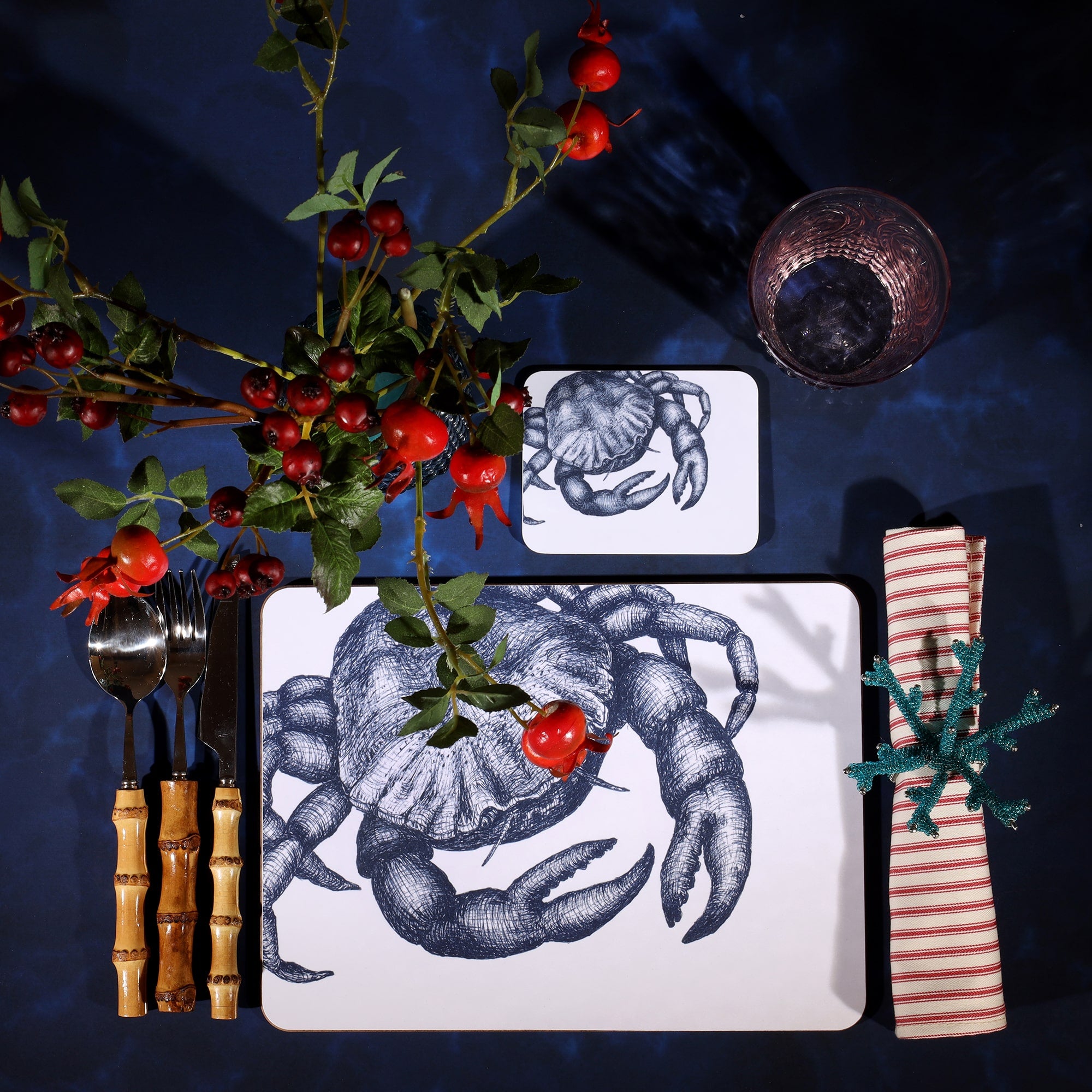 Crab Design in Navy on a white Coaster with a matching Placemat in a table setting on a dark table cloth.On the table is bamboo cutlery, stripe napkin and a glass with rosehips.