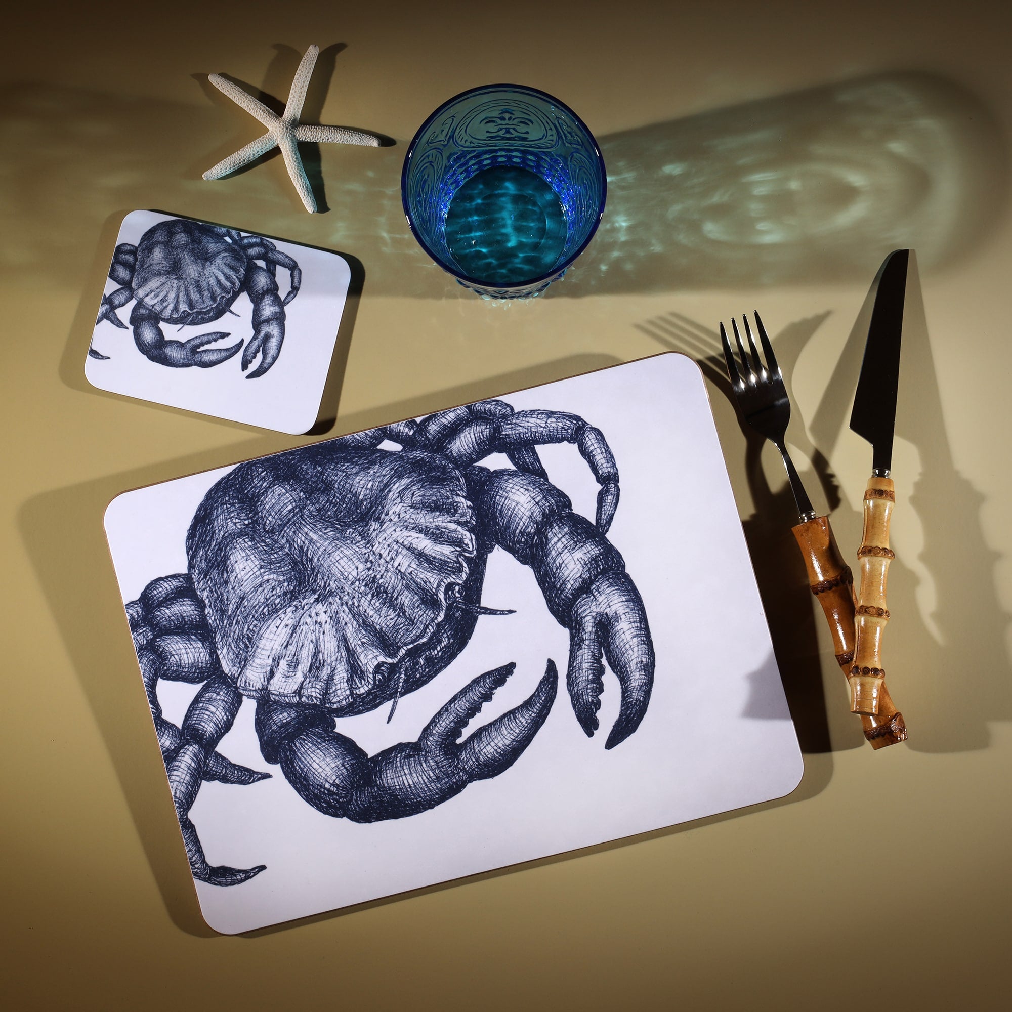 Crab Design in Navy on a white Coaster with a matching Placemat.On the table is a bamboo cutlery set and a blue coloured glass