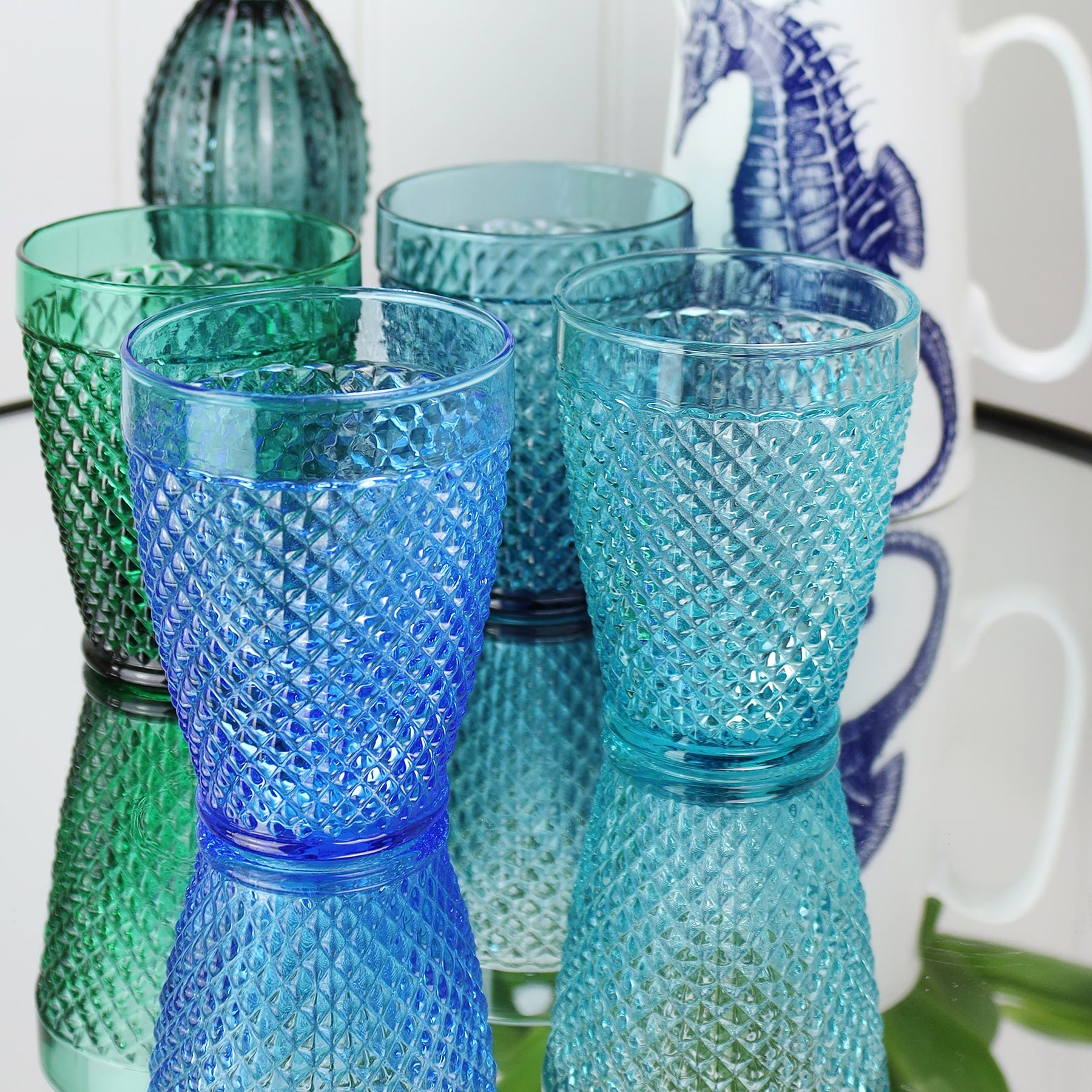 Lifestyle shot of four diamond cut tumblers on a mirror top table,in the background is a seahorse jug