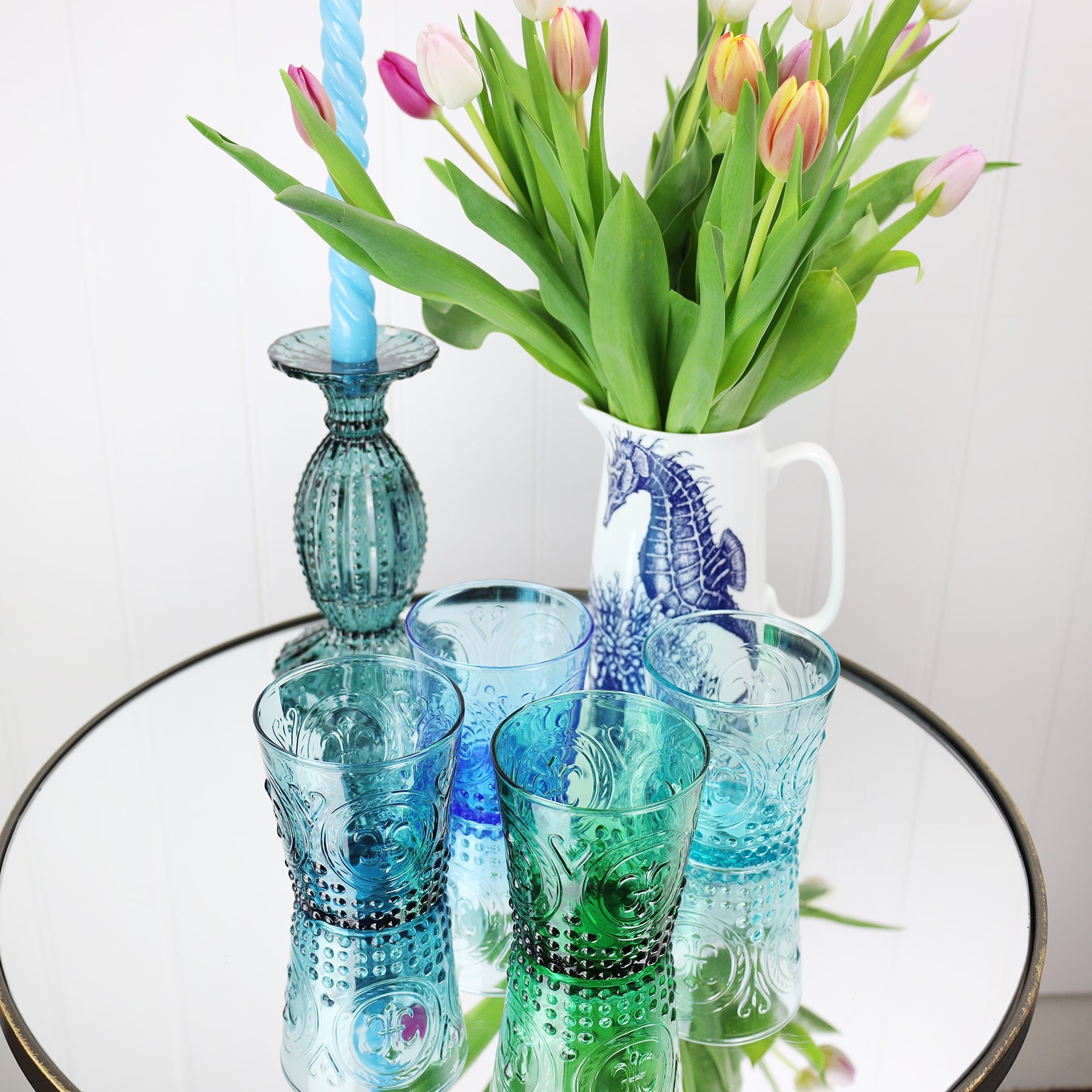 Teal Fleur de Lys Tumbler with other coloured glass tumblers on a mirror table with a seahorse jug filled with tulips