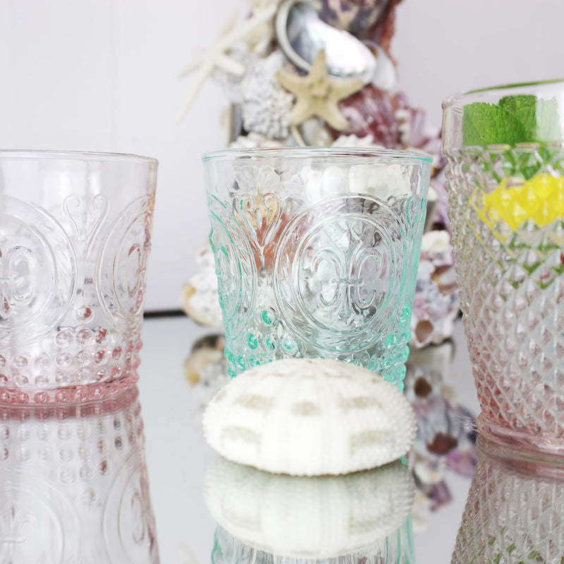 Close up of rose diamond cut tumbler on a mirror table with other coloured glasses, shell covered  candleholder in the background