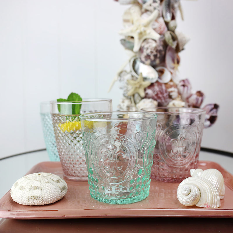 Aqua Fleur de Lys style glass tumbler on a tray with other glass tumblers with a shell covered candleholder in the background