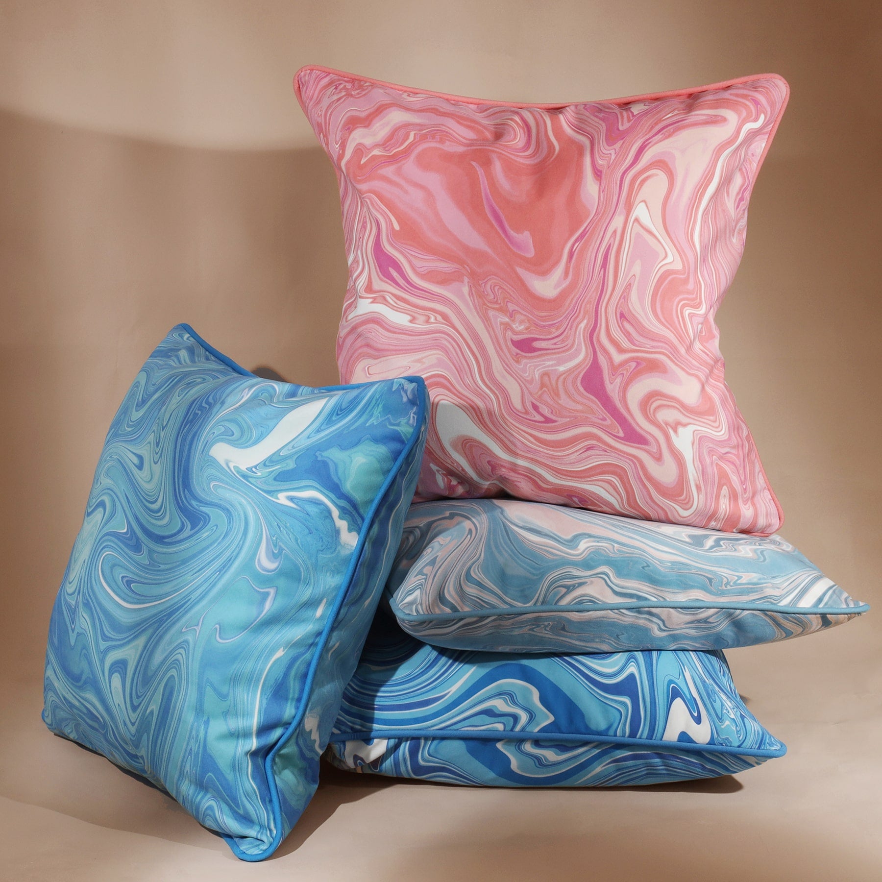 Marble,swirl cushion in Sundowner colours in pinks/whites and coral placed on top of a pile of similar designs in blues. greys and a brighter blue