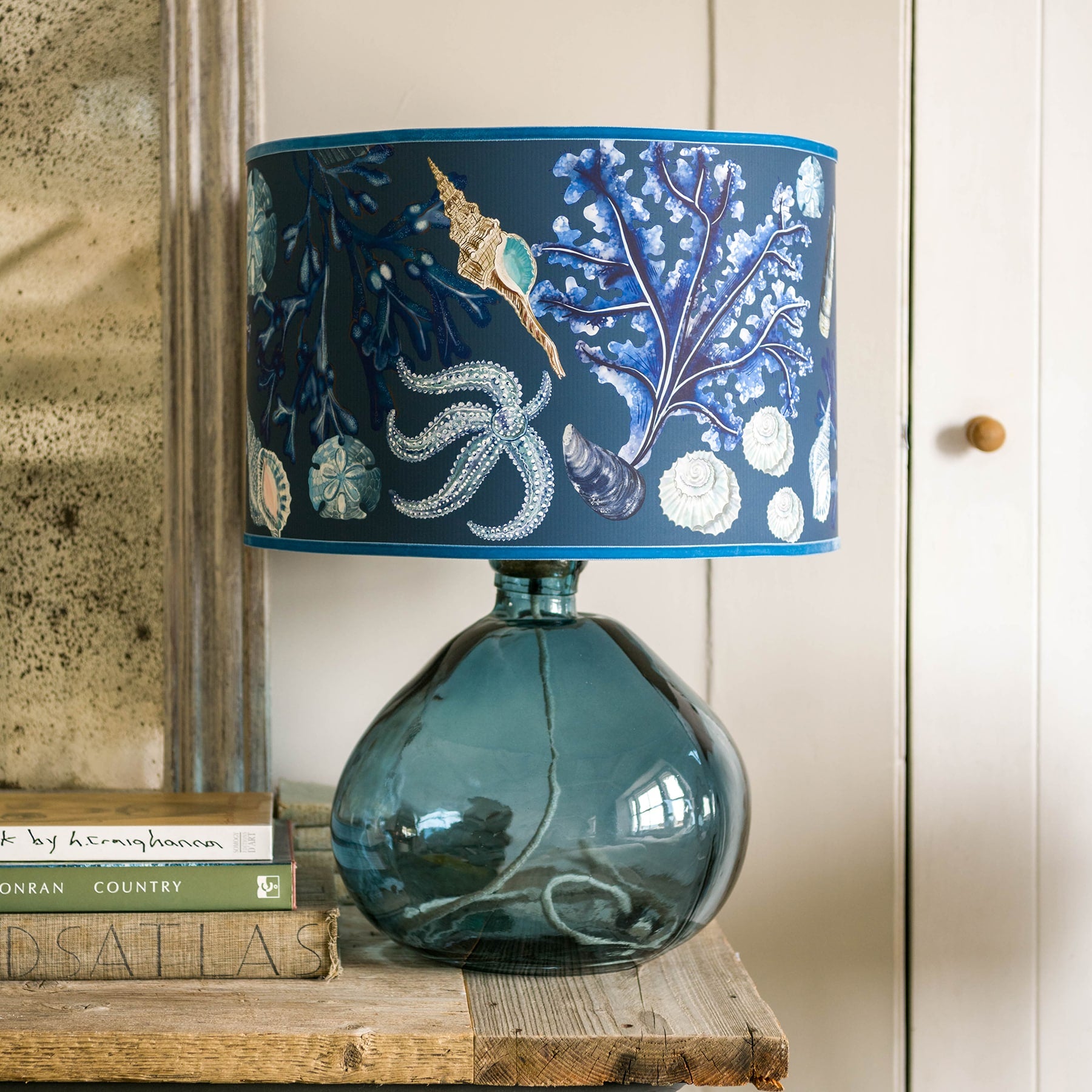 Rockpool Navy Shade With Shells,Seaweed,Sand dollars and Starfish Design in blues/browns with a blue trim on the edge of the shade on a glass lampbase.Its is placed on table next to a pile of books