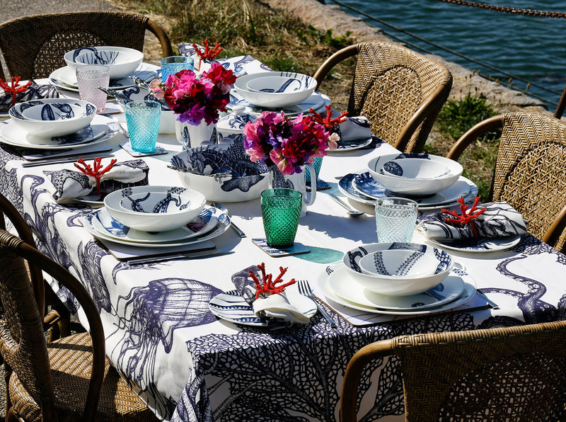 Table set by the sea with six place settings of our Bone China tableware on one of our tablecloths.On the table are two jugs with sweet peas and coloured glasses decorate the table.You can see the sea in the background