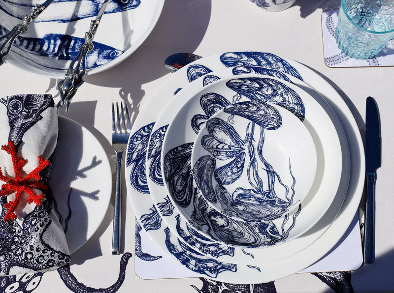 Outside Table setting with  Mussel & Oyster plates,stacked with the bowls on a placemat,next to a coaster with a blue glass.