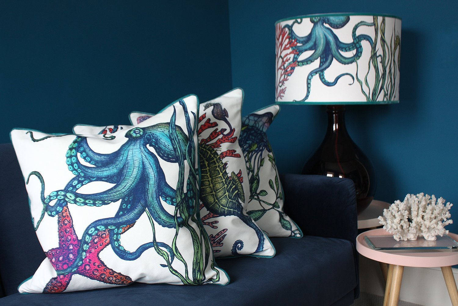 Rainbow Reef White Shade With Octopus,Seahorse,Starfish and Seaweed Design in bright colours on a navy glass lampbase next to a navy sofa covered in our Reef cushions.