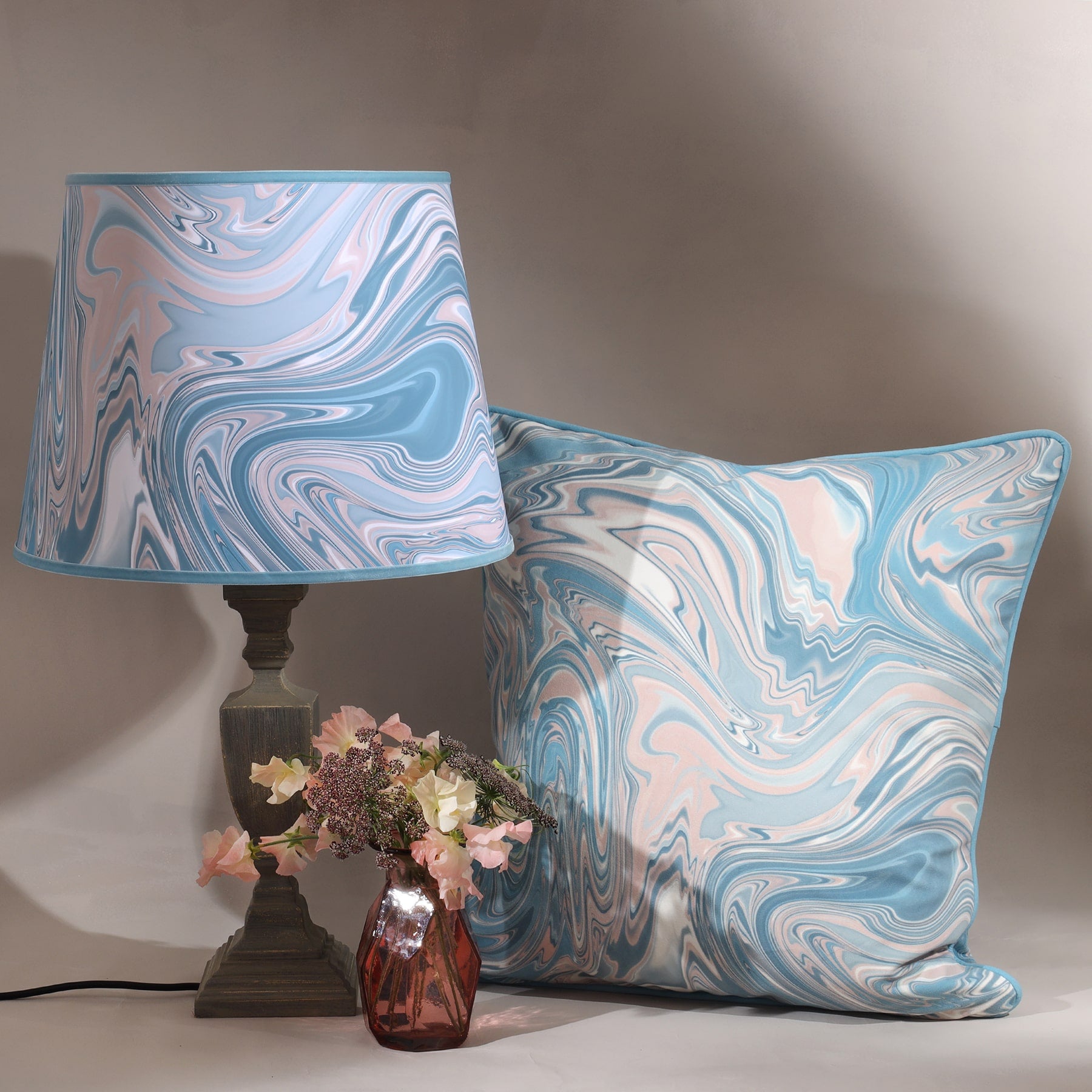 Ripple,marble design in beige/blues/greys on a lampshade on a wooden lampbase.Next to the base is a small glass vase with hydrandeas in,next to it is a matching cushion