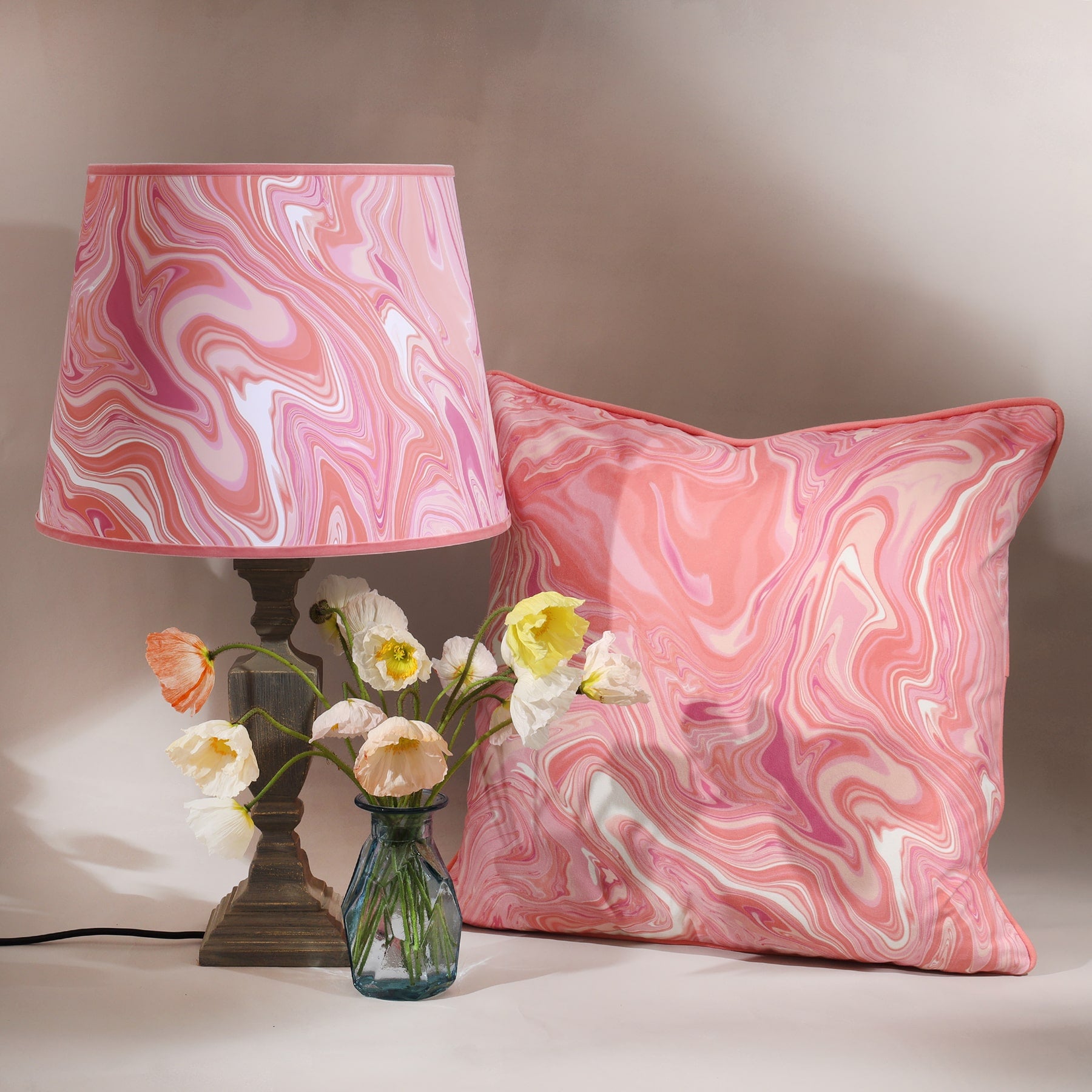 Marble,swirl cushion in Sundowner colours in pinks/whites and coral next to a matching lampshade and a base and a small vase of flowers