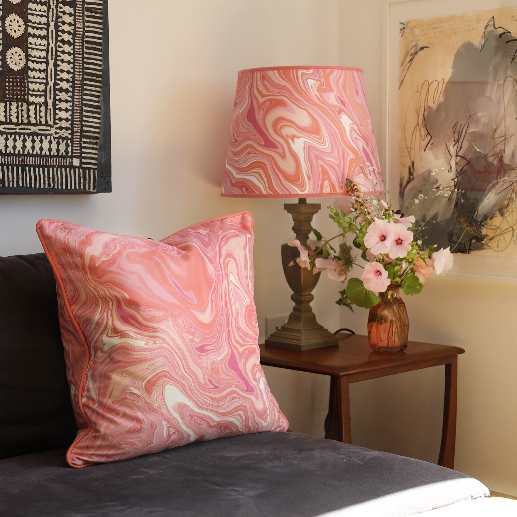 Marble,swirl cushion in Sundowner colours in pinks/whites and coral placed on a bed next to a matching lampshade and a vase on a table.