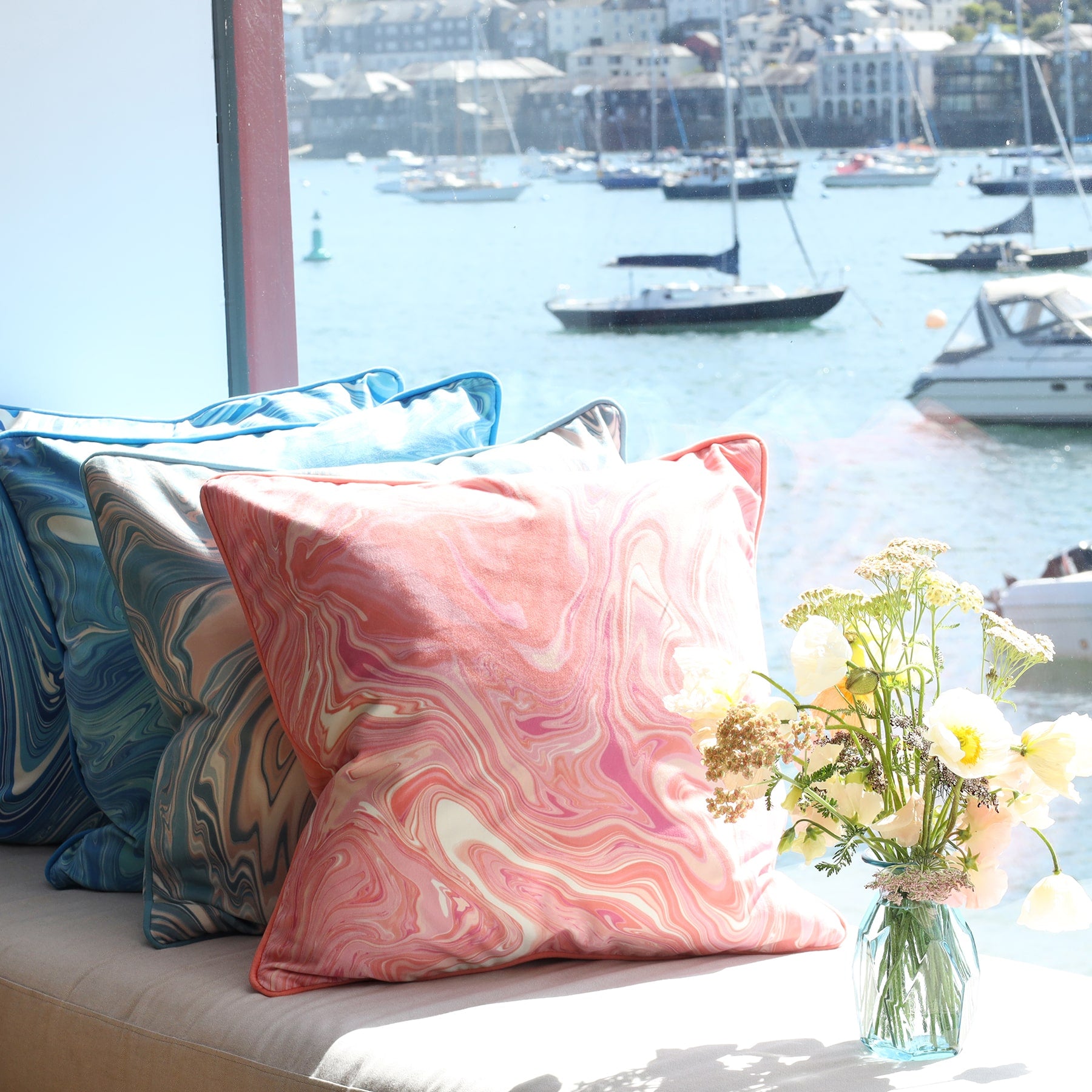 Marble,swirl cushion in Sundowner colours in pinks/whites and coral placed next to similar design in blues.All on a window seat with the sea and boats in the background