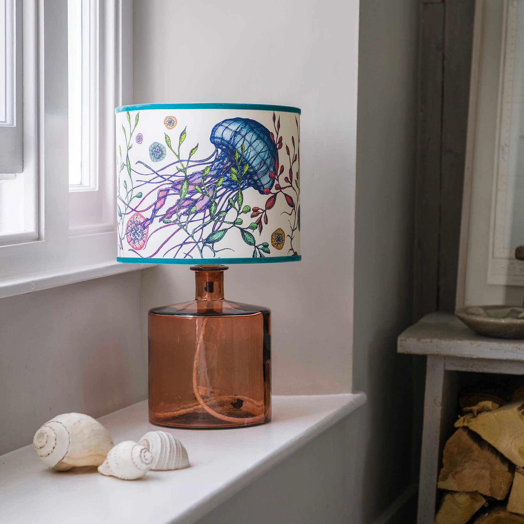 Canyons Reef White Shade With Jellyfish Design in bright colours on a pink glass lampbase placed on a window ledge.Next to the window is a table with firewood in.