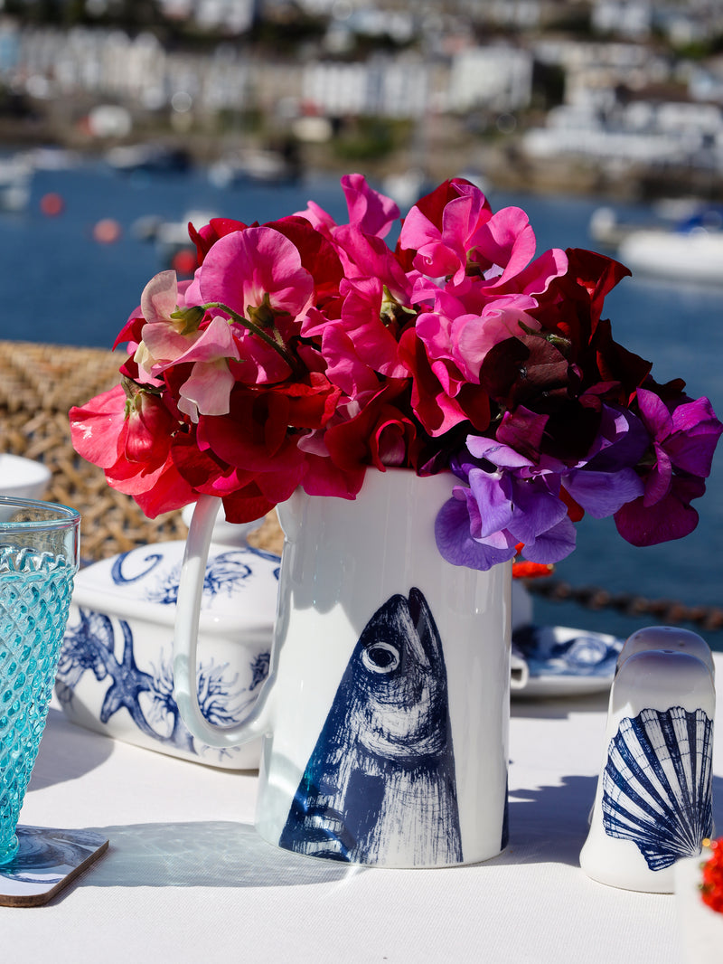 Bone China white Jug with hand drawn illustration of our Mackerel Heads in Navy.It holds pink sweetpeas and is on a outdoor table ,in the background os the harbour and you can see a butter dish and a salt and pepper set