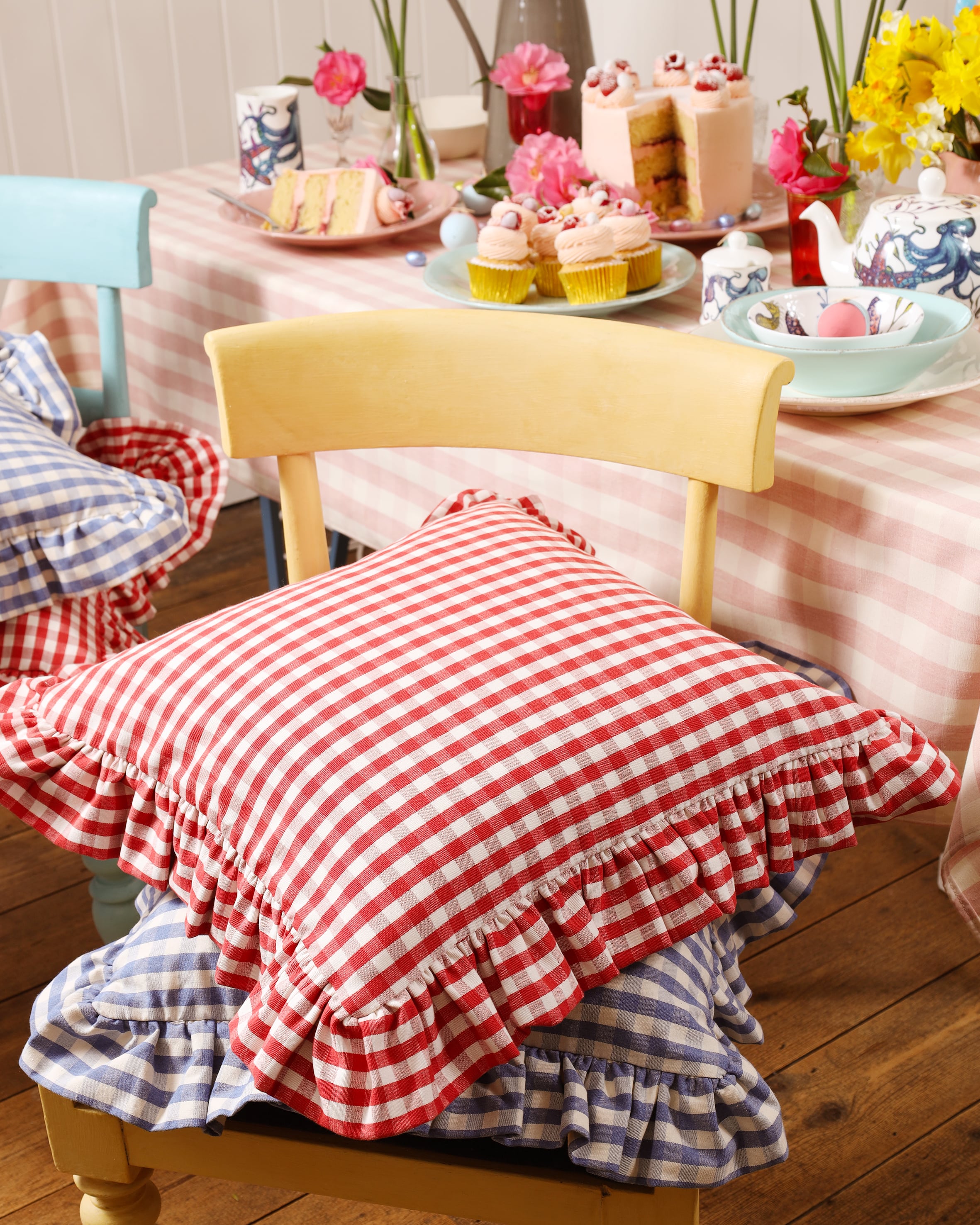 Red Gingham cushion on top of a blue gingham cushion placed on a yellow chair.Behind the chair you can see a table covered with a tablecloth and decorated with cupcakes and other table decorations.