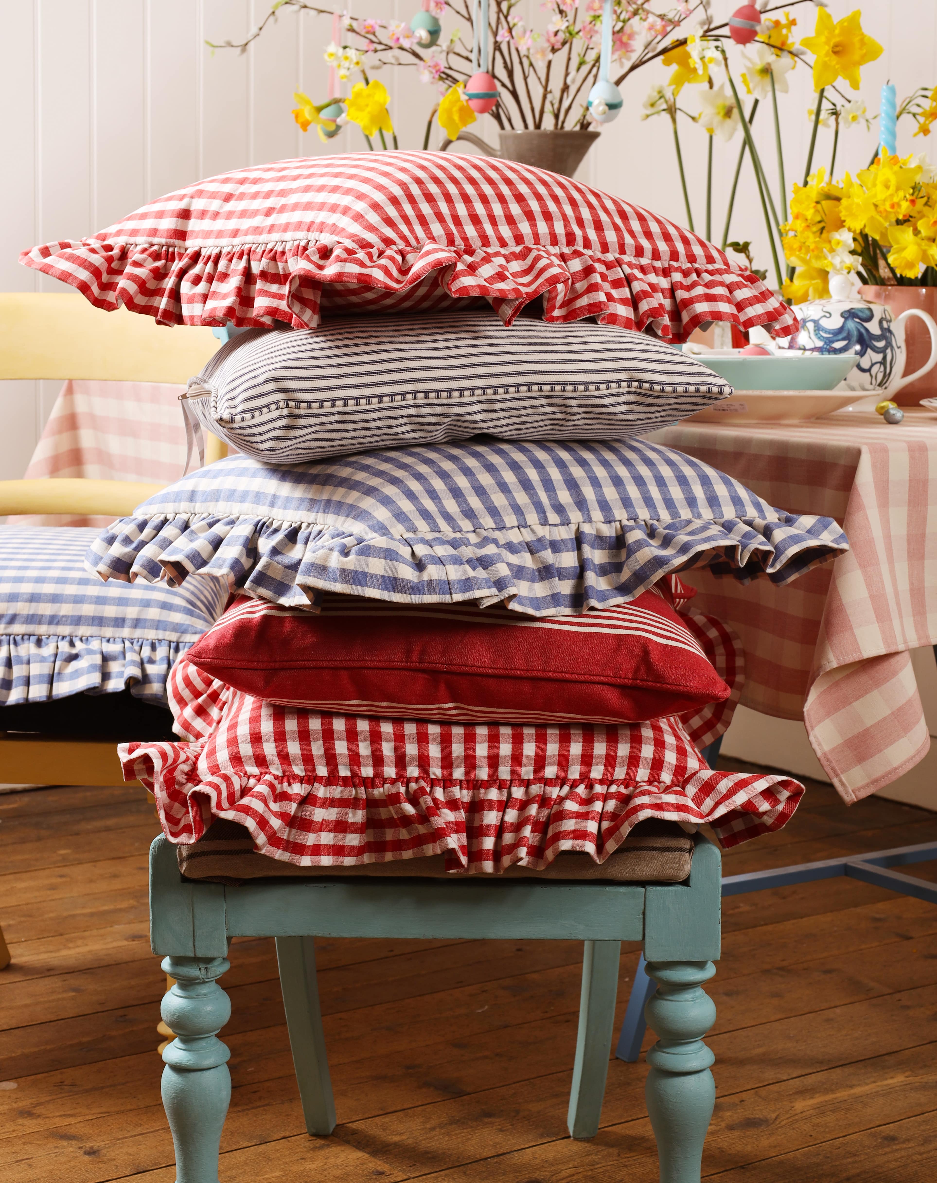 Red Gingham cushion on top of a blue gingham ,red striped,Navy striped pile of cushions in front of a table decorated with cakes and flowers.