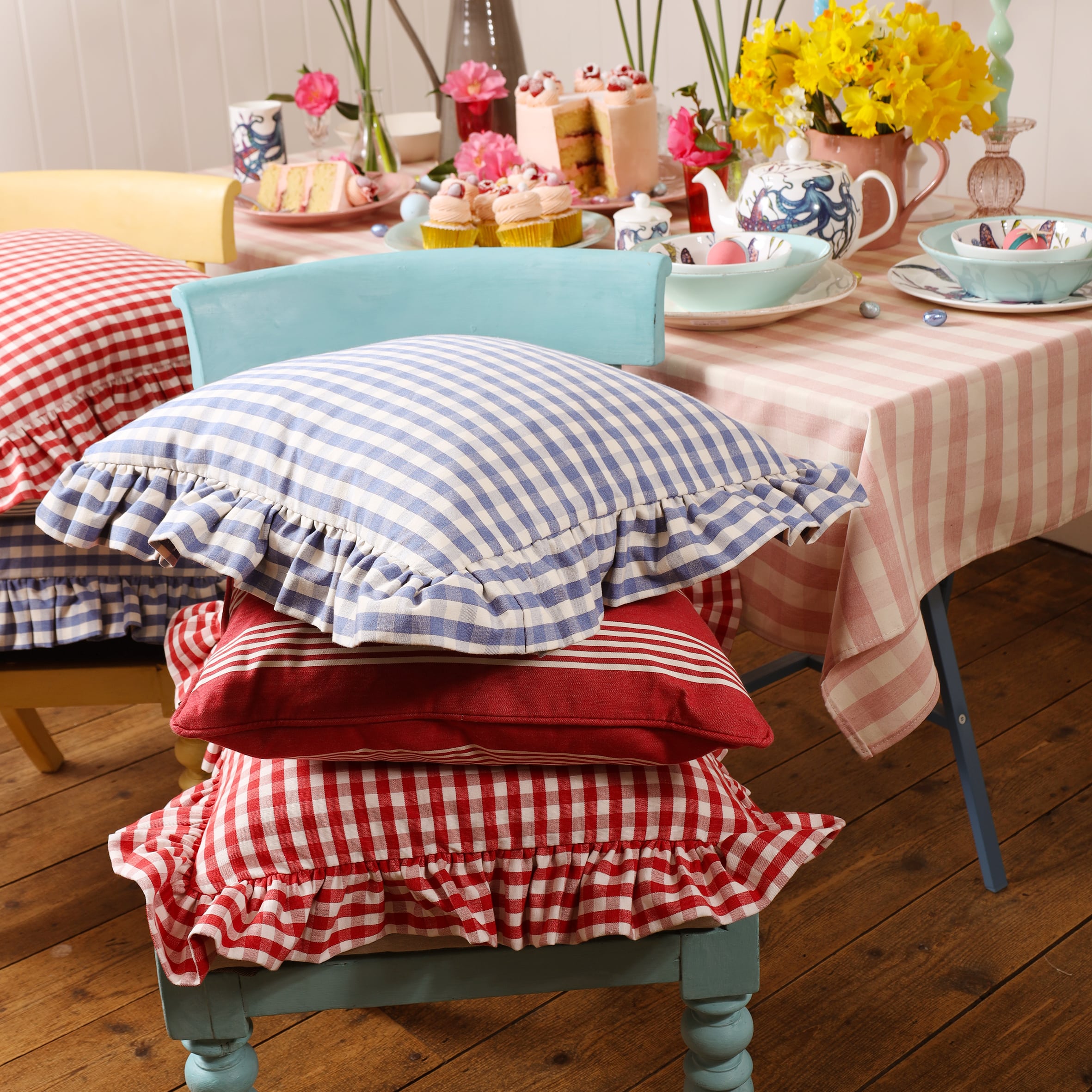 Blue and White Gingham cushion with a wide frill all around the edgeon other cushions including a Gingham cushion in red.Behind is a table dressed with a gingham tablecloth,on the tablecloth are lots of cupcakes,other cakes all placed on or around our Reef tableware.