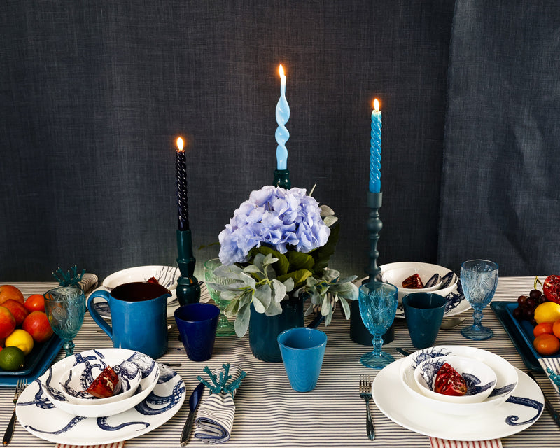 Side view of Bone China White plate with hand drawn illustrations of our Classic Octopus in Navy stacked with the dinner plate,pasta bowl and a cereal bowl with cutlery on a navy stripe tablecloth.On the table are different shades of blue candles/candleholders and blue drinking glasses