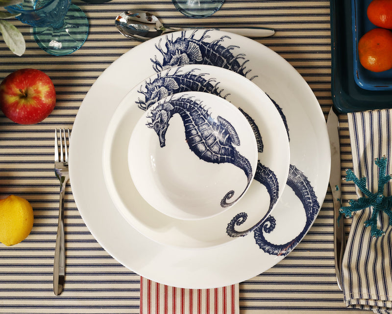 Aerial view of Bone China White plate with hand drawn illustrations of our Classic Seahorse in Navy stacked with the dinner plate,side plate and a bowl.Set in place setting with cutlery on a navy stripe tablecloth