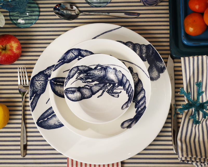 Aerial view of pasta bowl in Bone China in our Classic range in Navy and white in the Lobster design,placed on a matching dinner plate and has a smaller bowl placed inside.This is placed on a stripe tablecloth with various other tableware around the edge