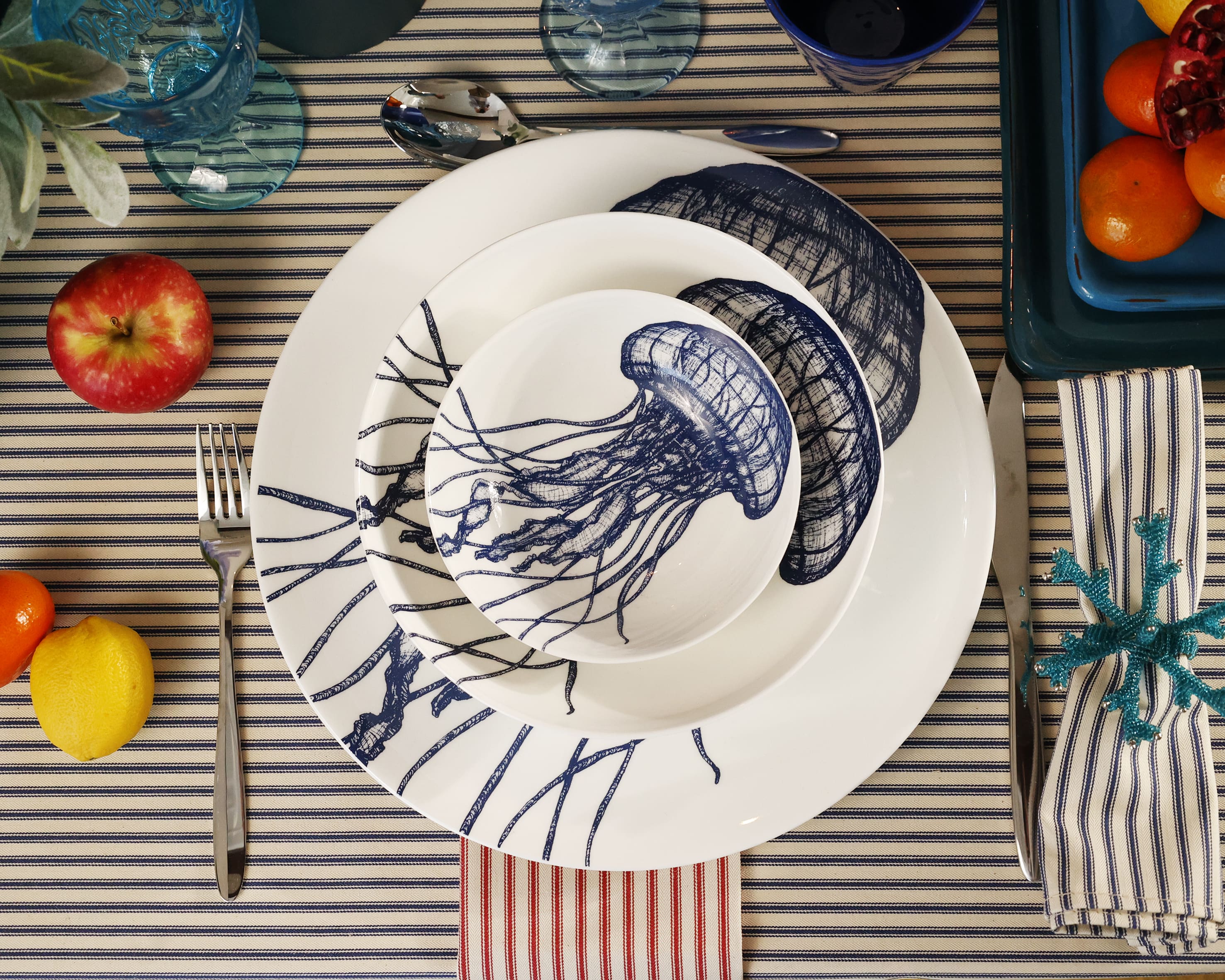 Aerial view of Bone China White plate with hand drawn illustrations of our Classic Jellyfish in Navy stacked with the dinner plate,side plate and a  bowl.Set in place setting with cutlery on a navy stripe tablecloth