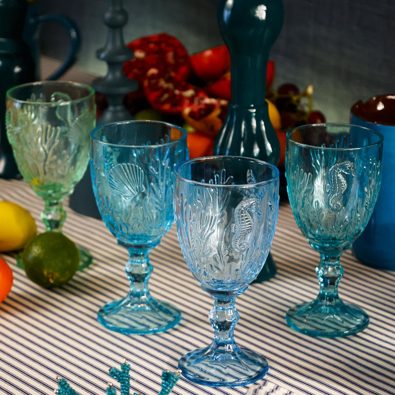 Set of 4 Underwater Goblets in four different shades of blues with embossed sea creatures in the glass, all placed on a table cloth.In the background you can see candle holders and other place settings