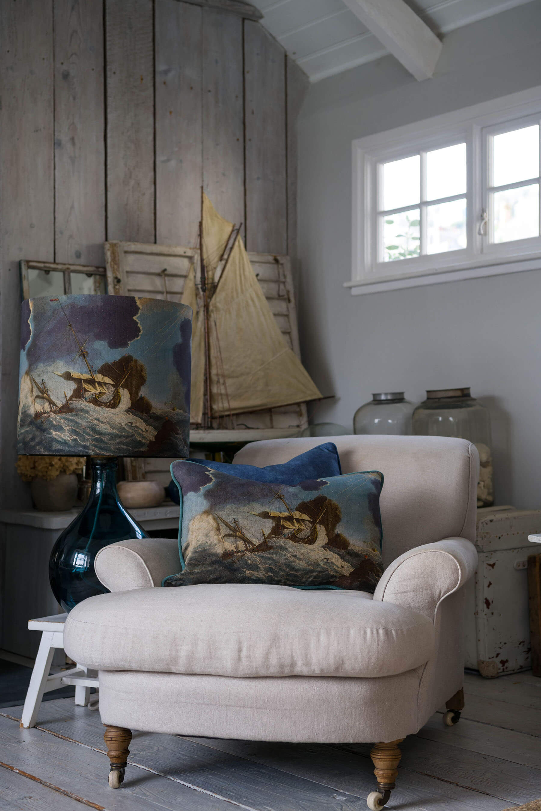 Shipwreck day cushion placed on a cream chair in front of a decorative sailing boat and other decorative items on a side table.Next to the chair is a matching lampshade on a petrol glass lampbase.