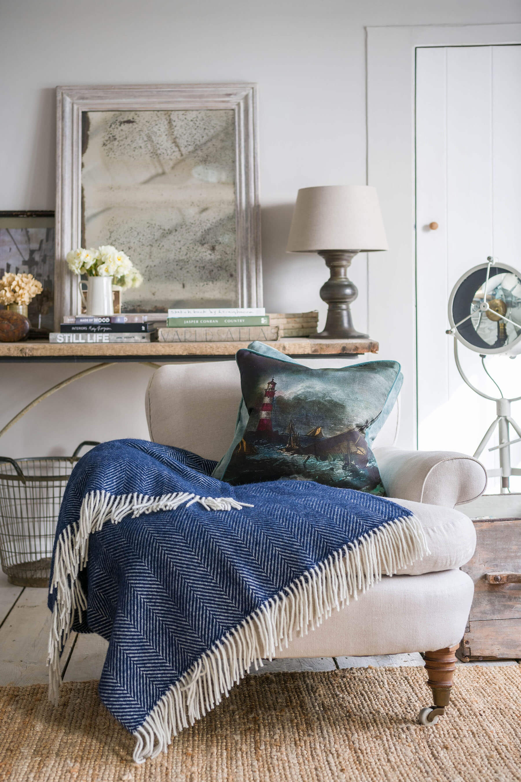 Lighthouse cushion placed on a cream chair over a navy throw.In the background is a table with books and distressed mirror and flowers on.Behind the chair is a door with a upstanding fan in front of it.