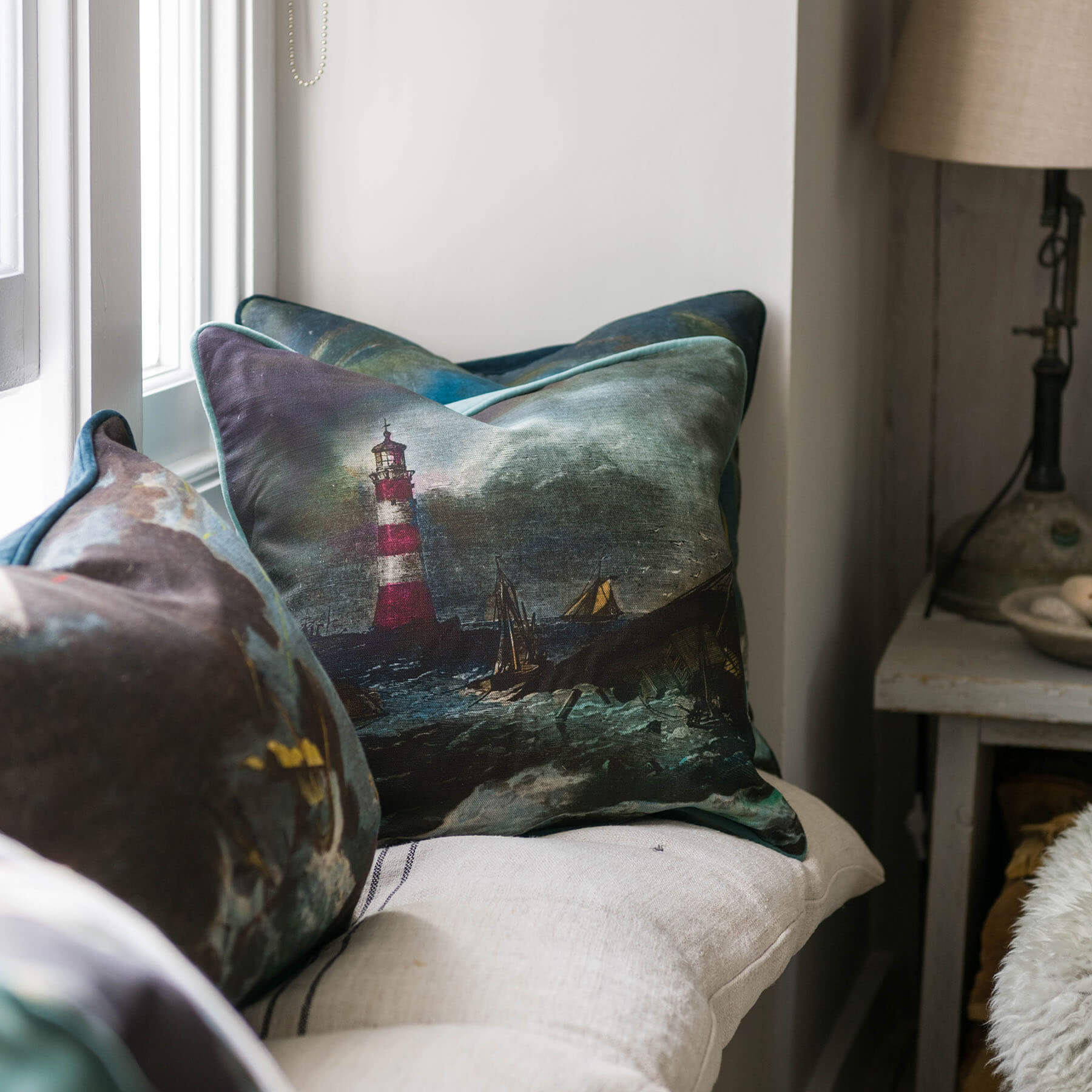 Lighthouse Cushion cover placed on a window seat next to other  shipwreck cushions,in the background you can see a table with a lamp on