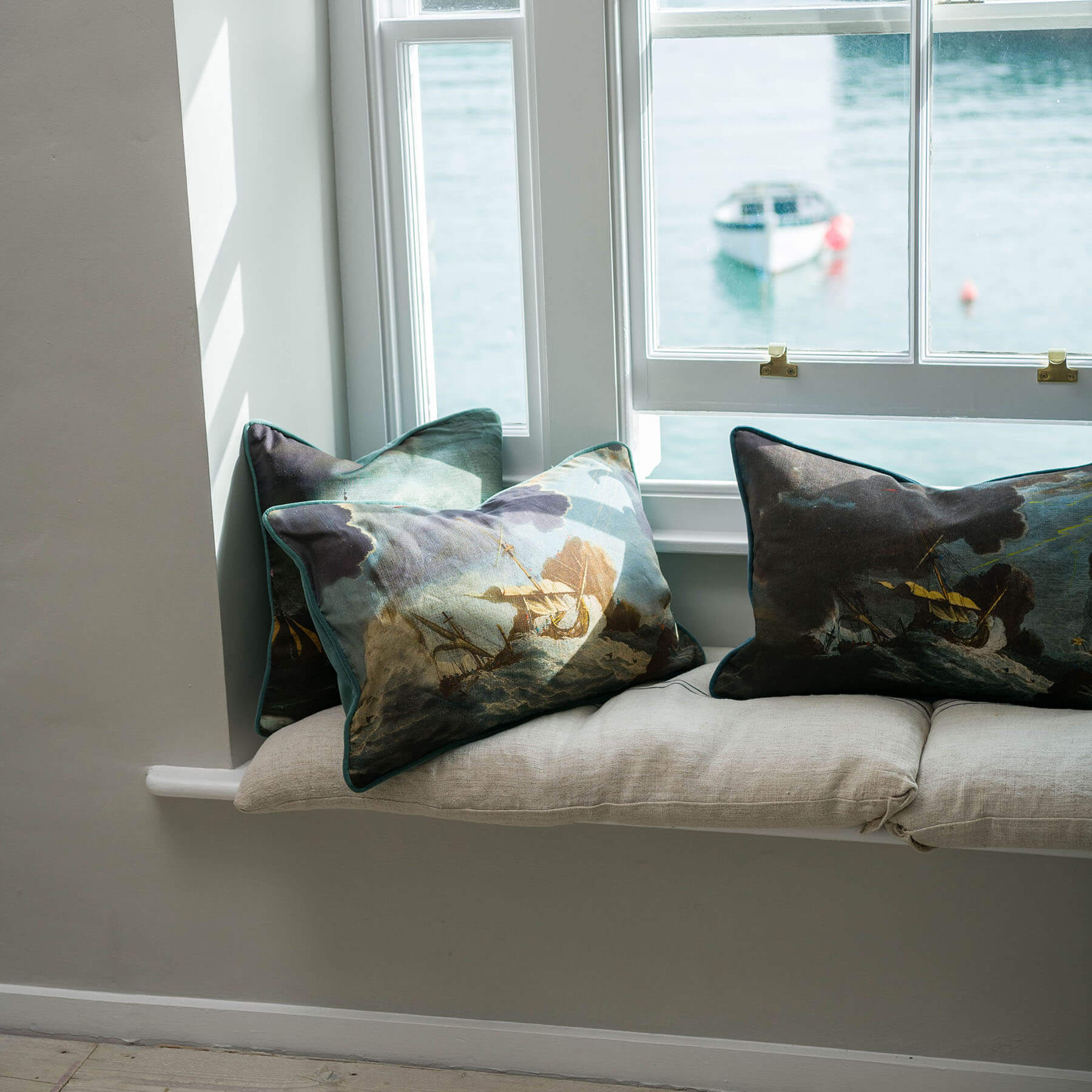 Shipwreck day cushion placed on a windowsill with other shipwreck cushions.You can see the sea outside and a small boat.