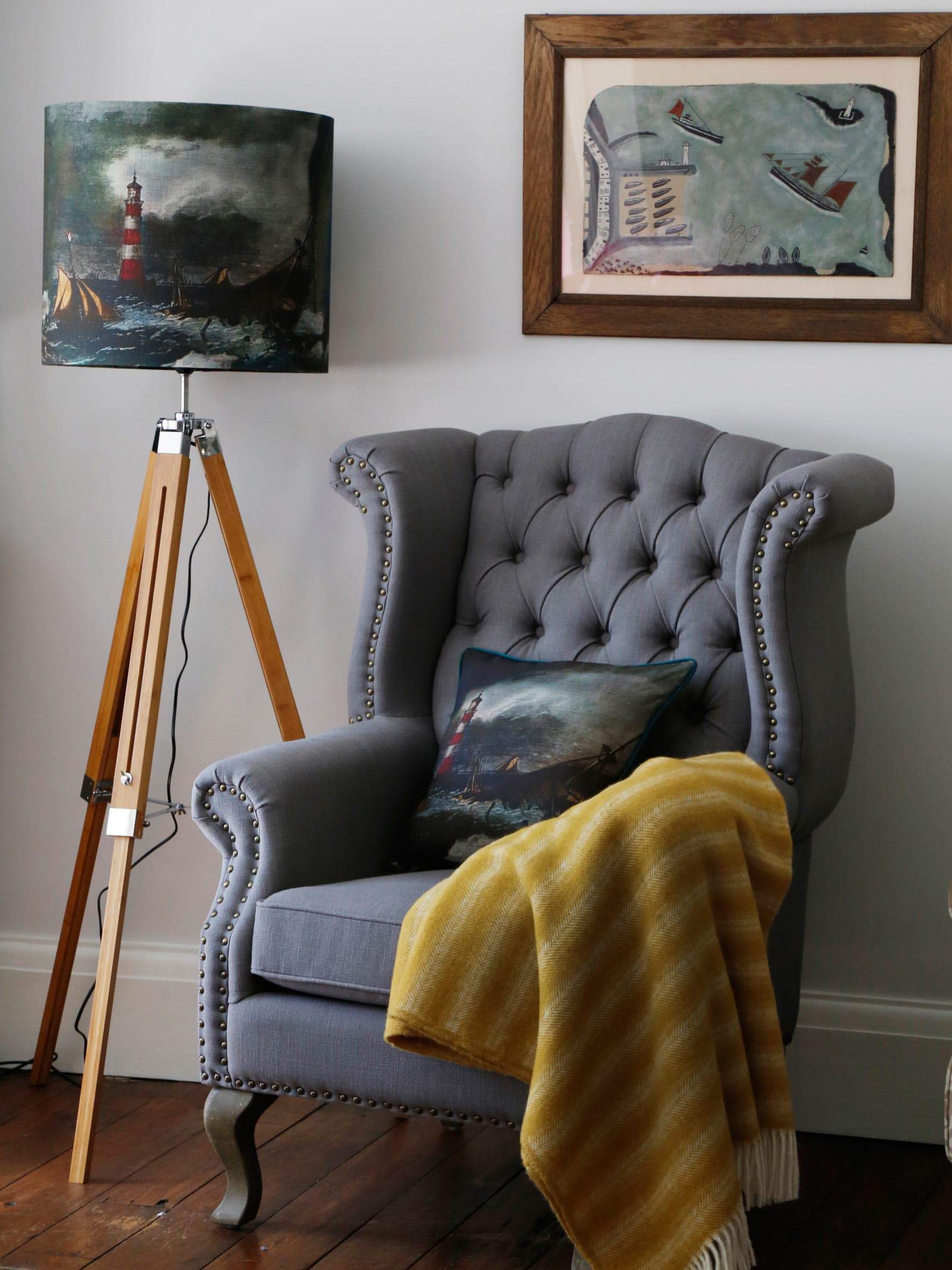 Lighthouse Cushion placed on a Grey Wingbacked chair with a mustard throw draped over the arm.The chair is next to a Tripod with a matching Lampshade.Behind on the wall is an Alfred Wallis Style painting