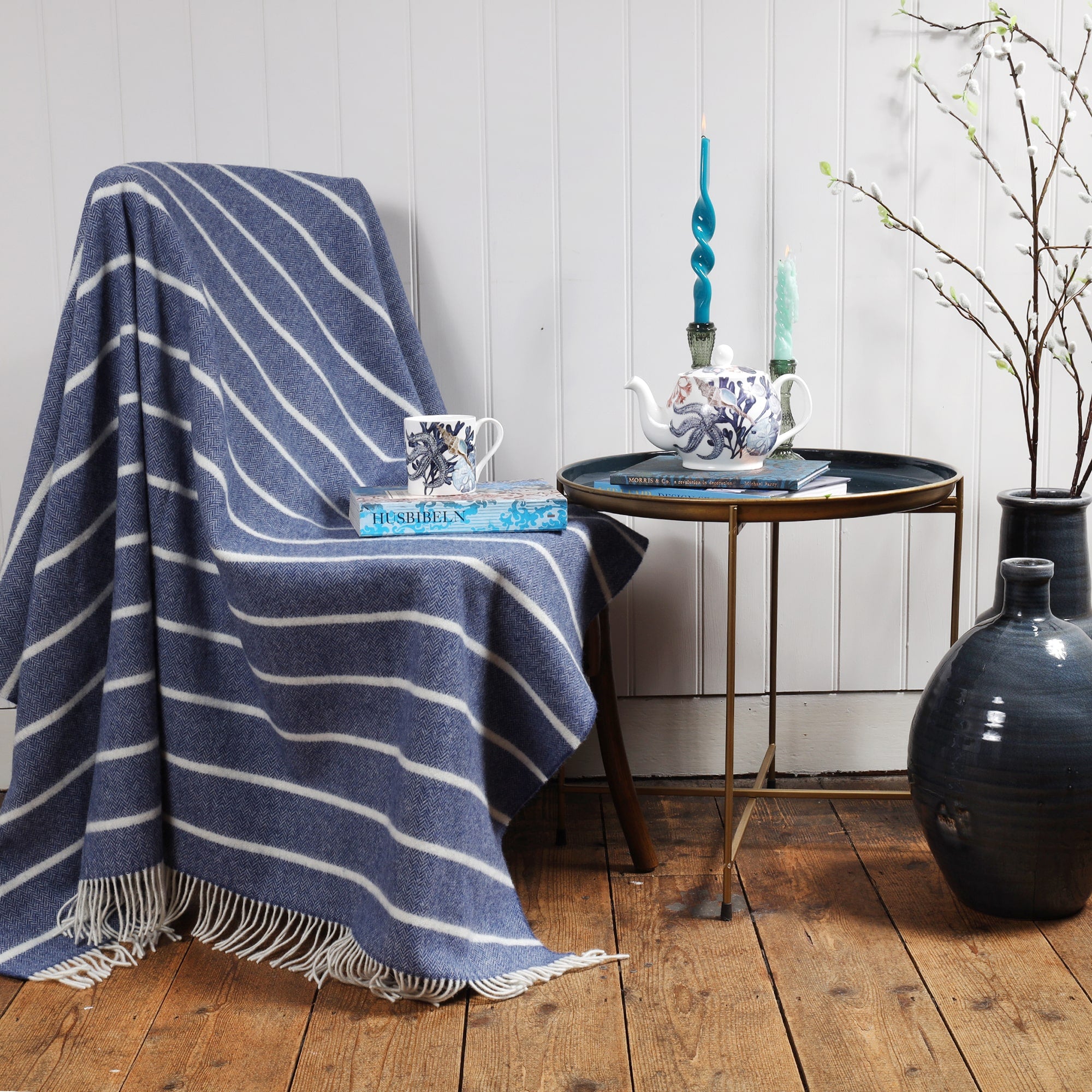 Lambswool Denim and White striped throw draped over a chair.Placed on the chair is a book and a mug in Beachcomber design,next to the chair is a table with books and a beachcomber Teapot and candles and candle holders.Next to the table are large navy vases with catkins coming out of them