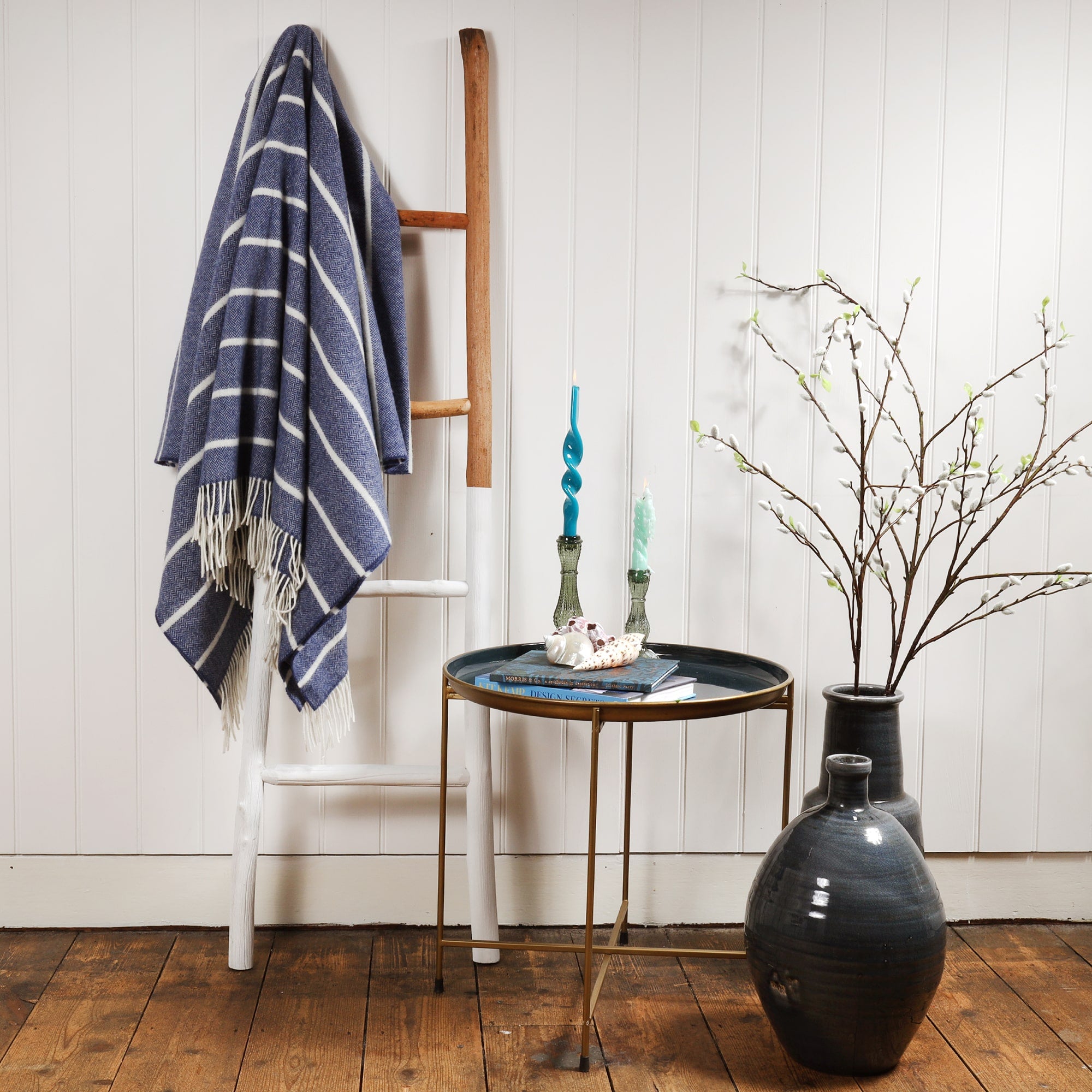 Lambswool Denim and White Stripe throw draped over a ladder against a wall.Next to the ladder is a table with books and a beachcomber Teapot and candles and candle holders.Next to the table are large navy vases with catkins coming out of them