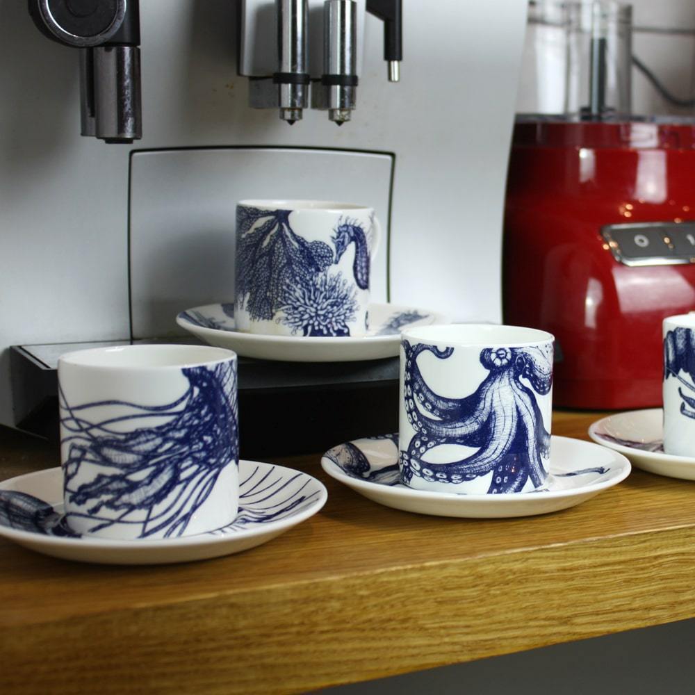 Side view of Four Bone China white espresso cups in hand drawn illustrations in our Octopus,Seahorse,Jellyfish and the Lobster designs in Navy with matching saucers in front of large coffee machine and a red food processor 