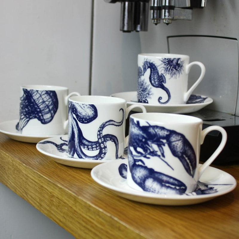 Side view of Four Bone China white espresso cups in hand drawn illustrations in our Octopus,Seahorse,Jellyfish and the Lobster designs in Navy with matching saucers in front of large coffee machine 