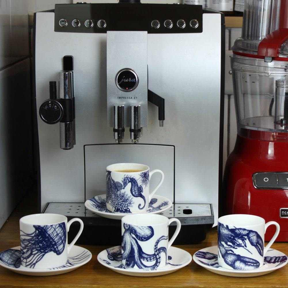 Four Bone China white espresso cups in hand drawn illustrations in our Octopus,Seahorse,Jellyfish and the Lobster designs in Navy with matching saucers in front of large coffee machine next to a red food processor
