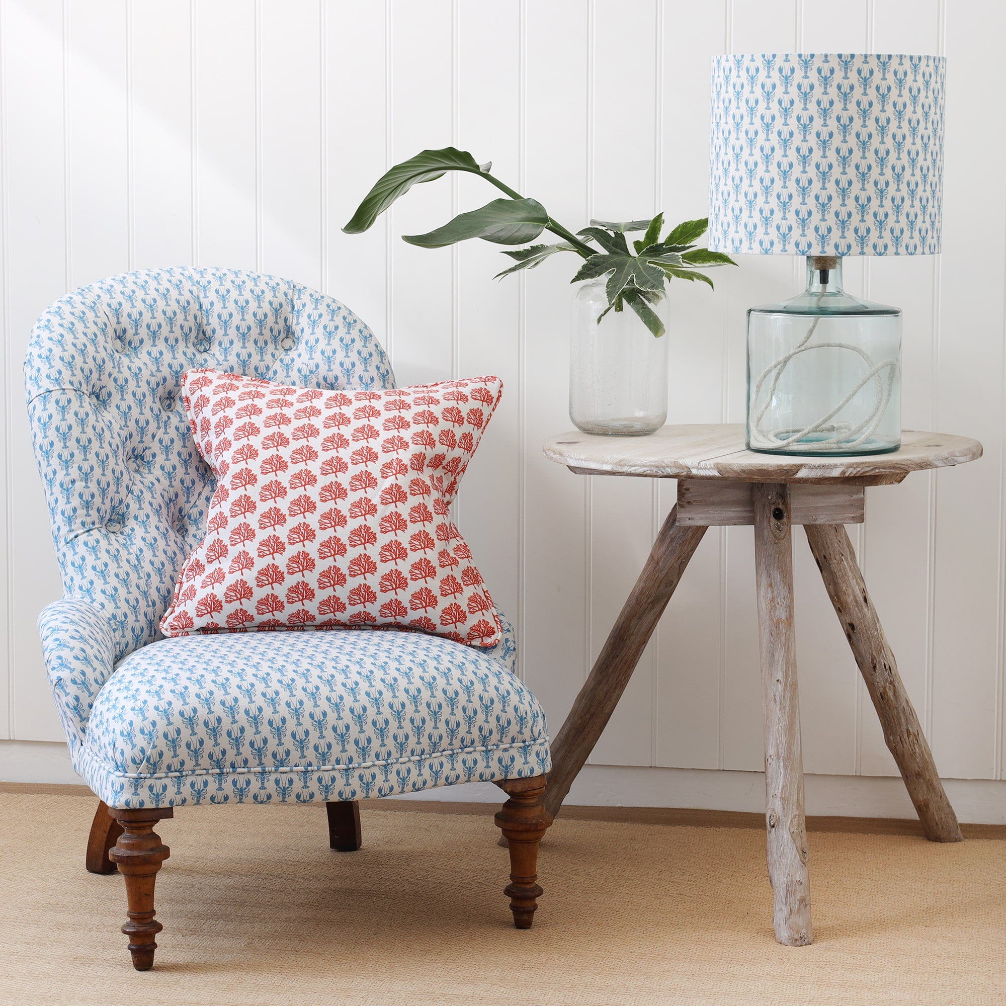 Off white mini coral cushion placed on a chair covered in a mini lobster print in cerulean Blue.next to the chair is a Tripod table with a glass lampbase with a matching mini lobster lampshade.