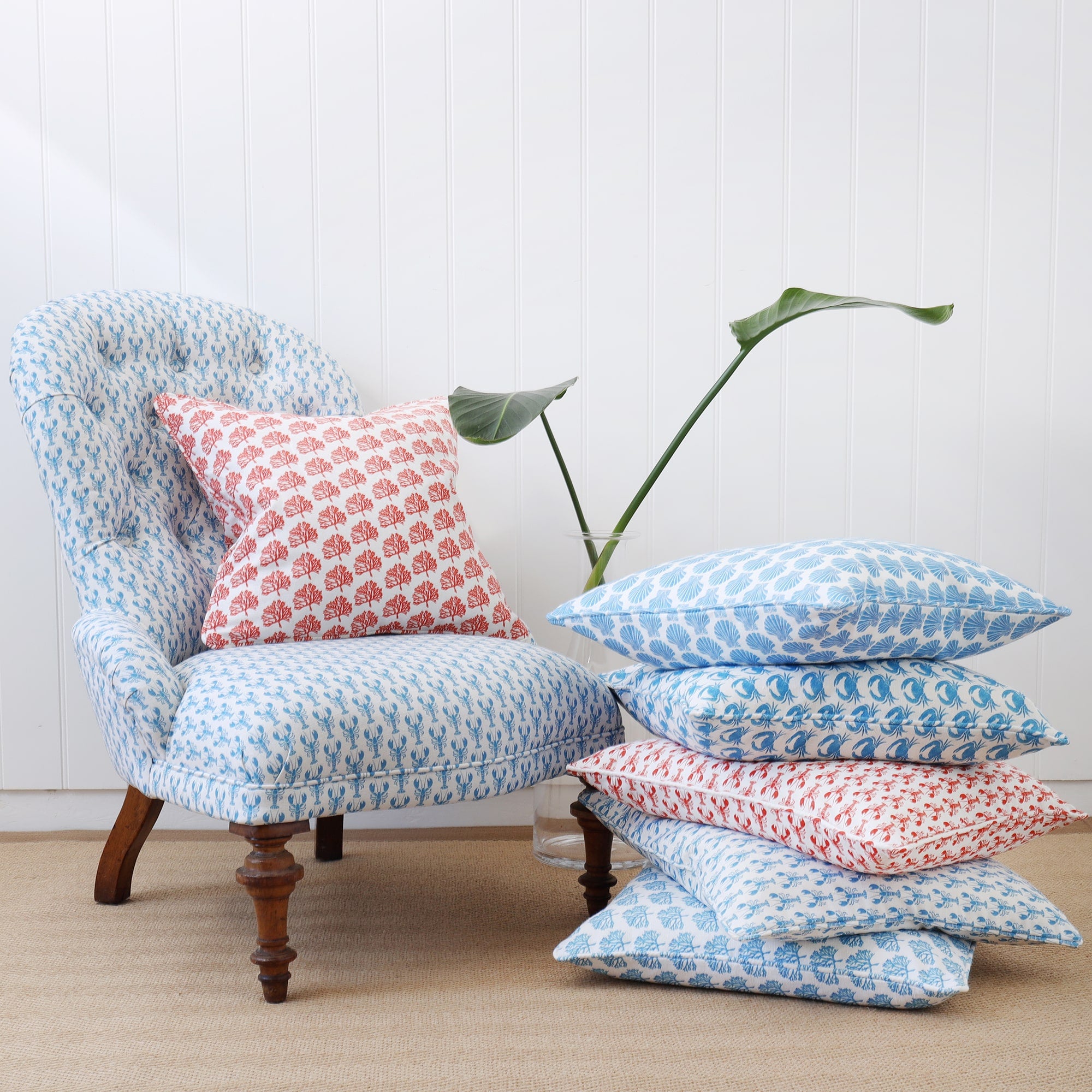 Off white mini coral cushion placed on a chair covered in a mini lobster print in cerulean Blue.next to the chair is a Tripod table with a glass lampbase with a matching mini lobster lampshade. Next to the chair is a pile of cushions with various mini prints on in various colours.