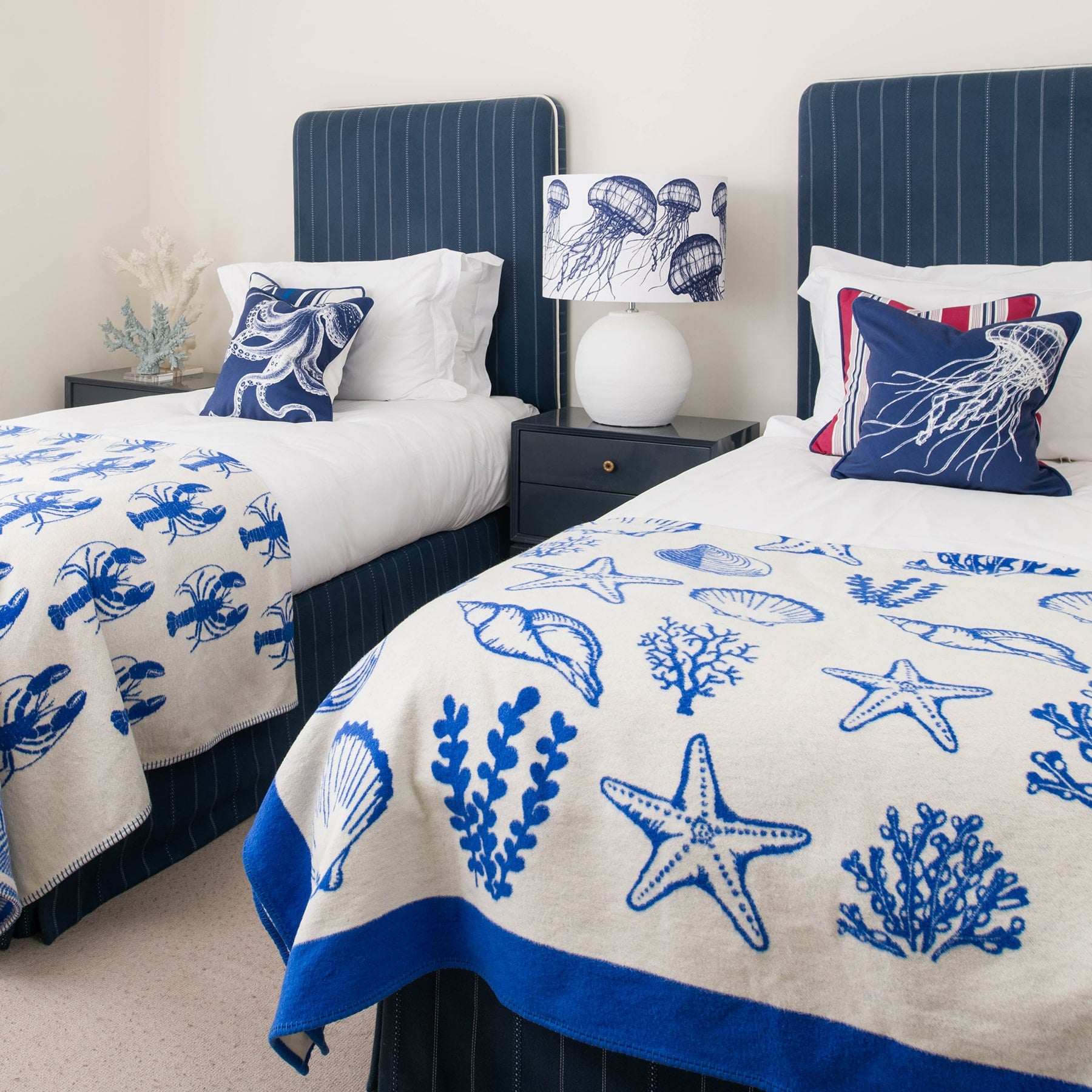 Our Classic Navy jellyfish design on a white background on a white lampbase on a side table between two single beds.On the beds are classic Navy and White cushions and our reversible throws are draped over the ends of the beds.