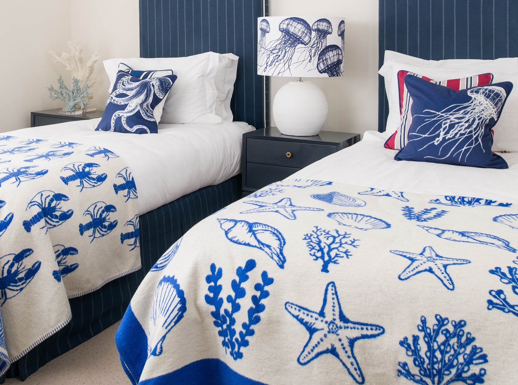 Our Classic Navy jellyfish design on a white background on a white lampbase on a side table between two single beds.On the beds are classic Navy and White cushions and our reversible throws are draped over the ends of the beds.