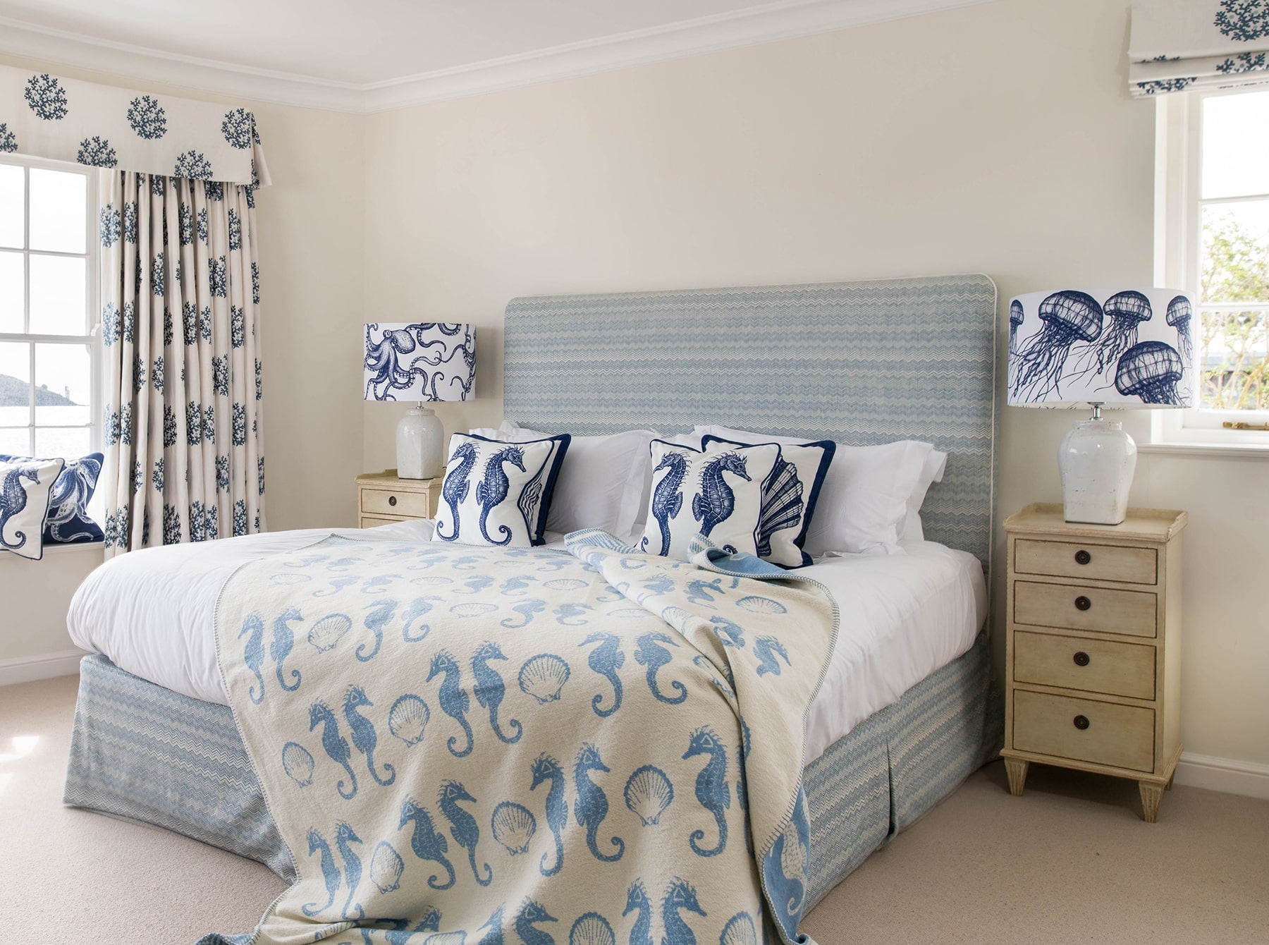Our Classic Navy jellyfish lampshade on either side of a double bed.On the bed are classic seahorse and scallop cushions on a soft blue seahorse reversible throw.
