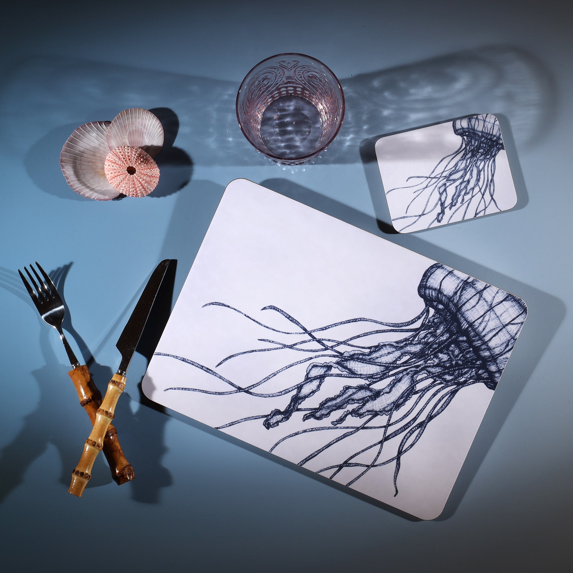 Jellyfish Design in Navy on a white Coaster with a matching Placemat.On the table is a bamboo cutlery set and a blue coloured glass