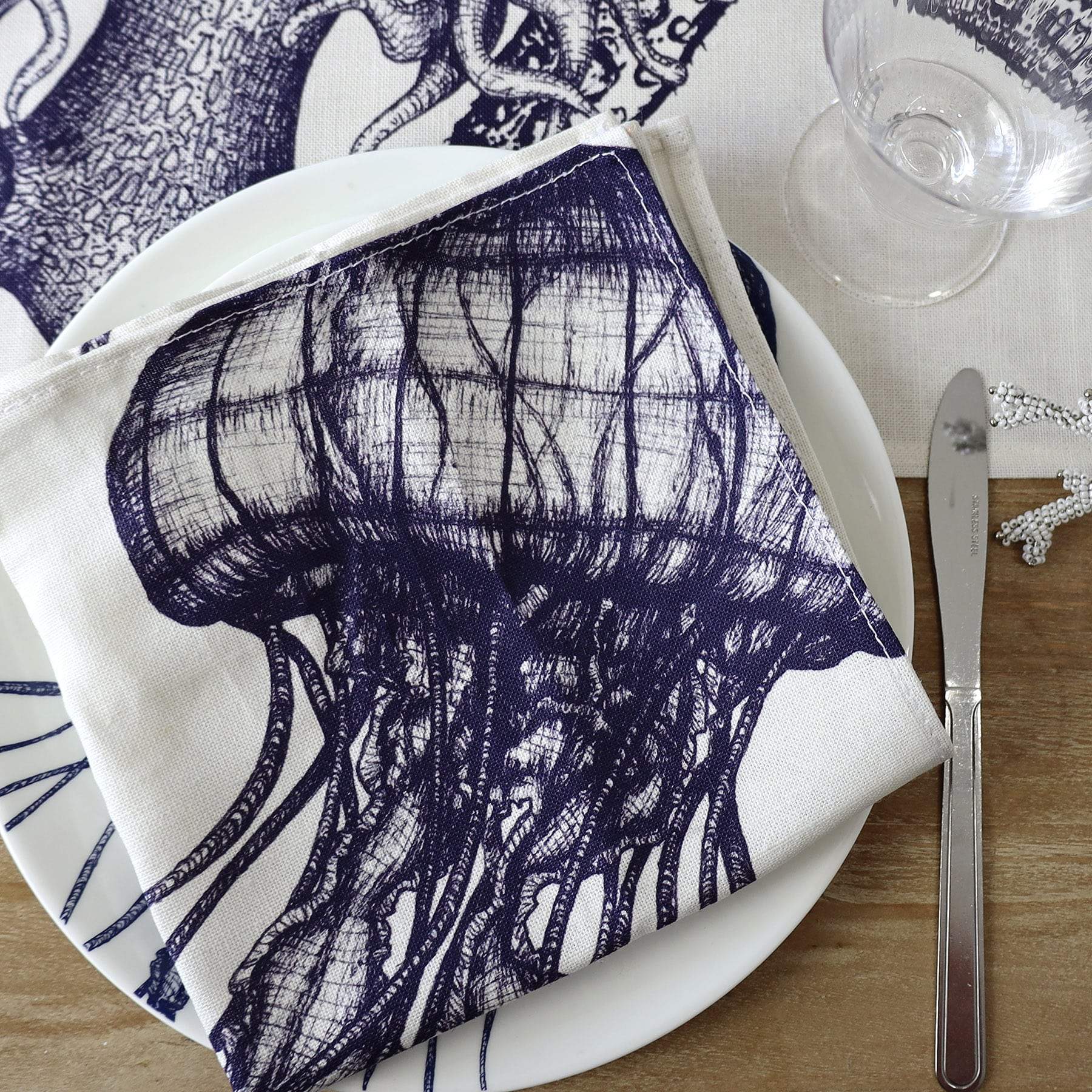 Blue & white linen napkins with seahorse, lobster, octopus and jellyfish hand drawn illustrations,set of four,close up of the Jellyfish one placed on an Jellyfish dinner plate.All placed on a sea creature tablecloth