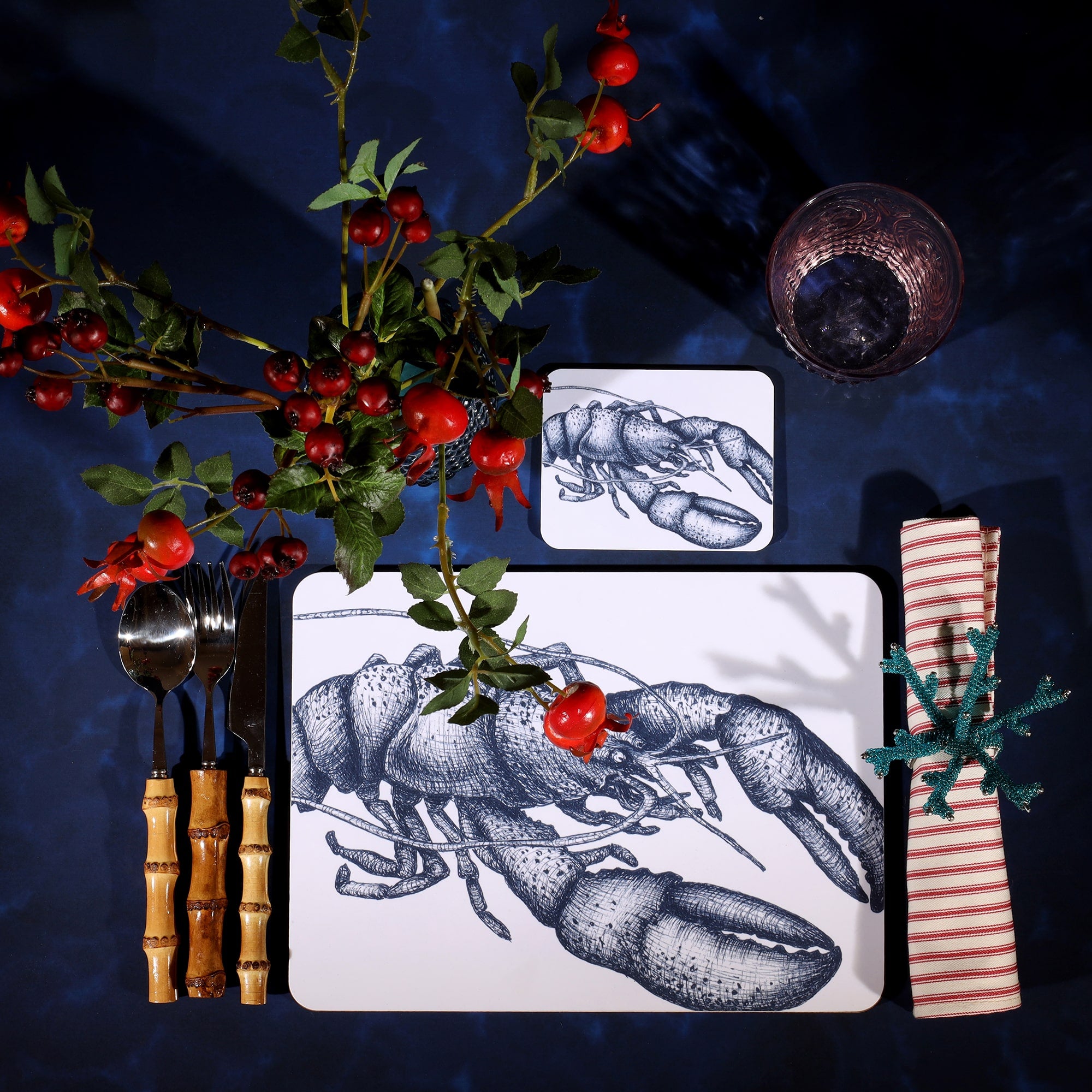 Lobster Design in Navy on a white Coaster with a matching Placemat in a table setting on a dark table cloth.On the table is bamboo cutlery, stripe napkin and a glass with rosehips.