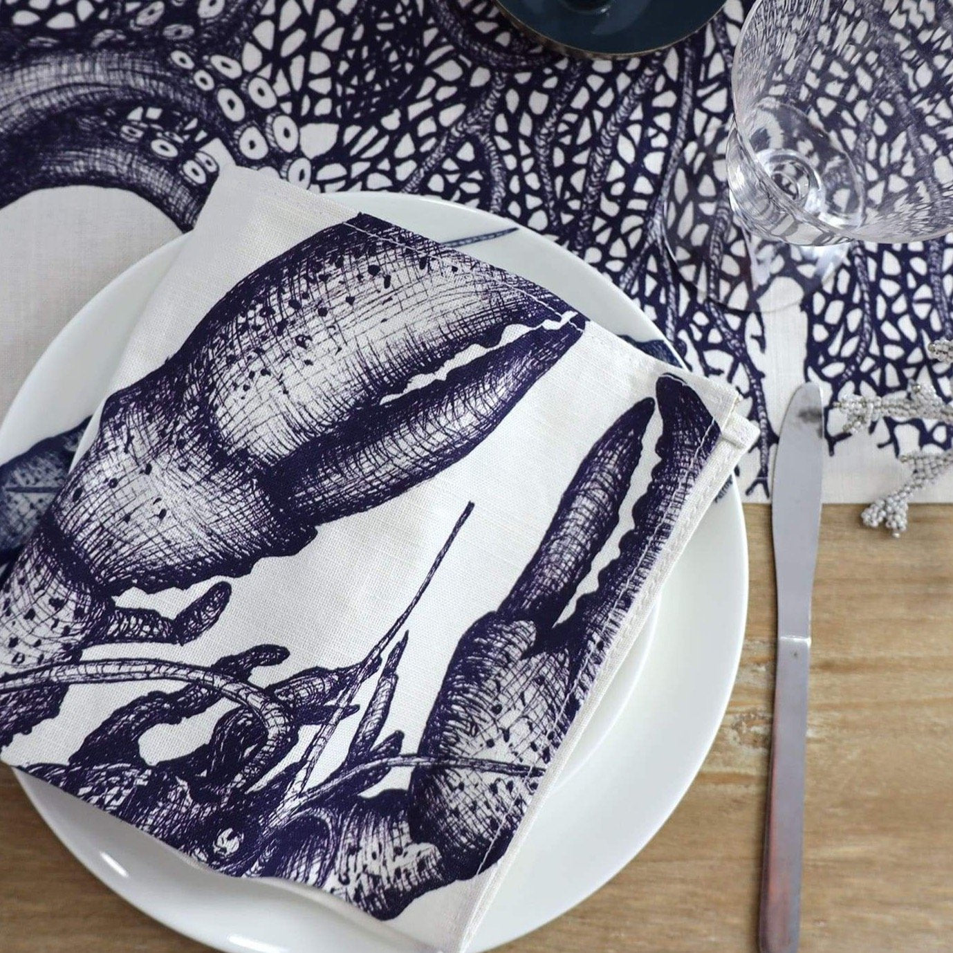 Blue & white linen napkins with seahorse, lobster, octopus and jellyfish hand drawn illustrations,set of four,close up of the Lobster one placed on a Lobster dinner plate.All placed on a sea creature table cloth