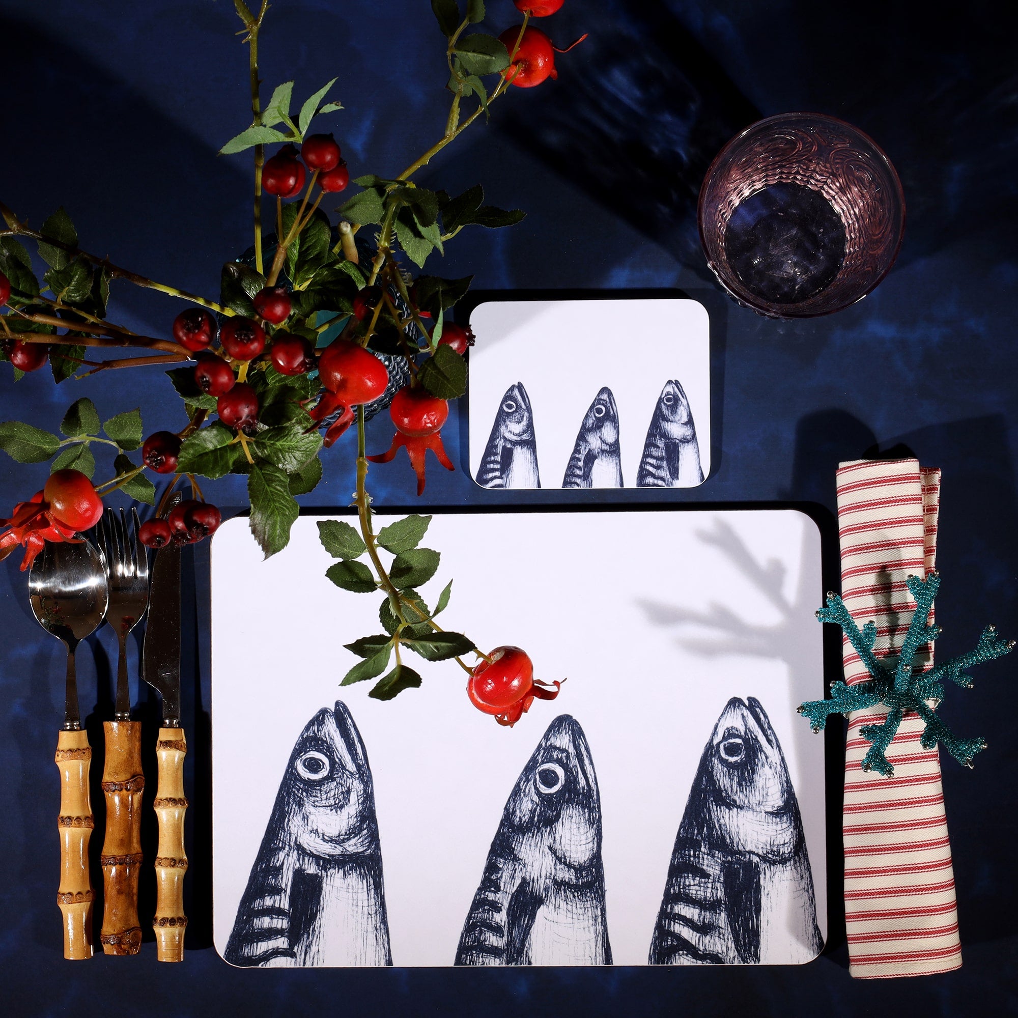 Mackerel heads Design in Navy on a white Coaster with a matching Placemat in a table setting on a dark table cloth.On the table is bamboo cutlery, stripe napkin and a glass with rosehips.
