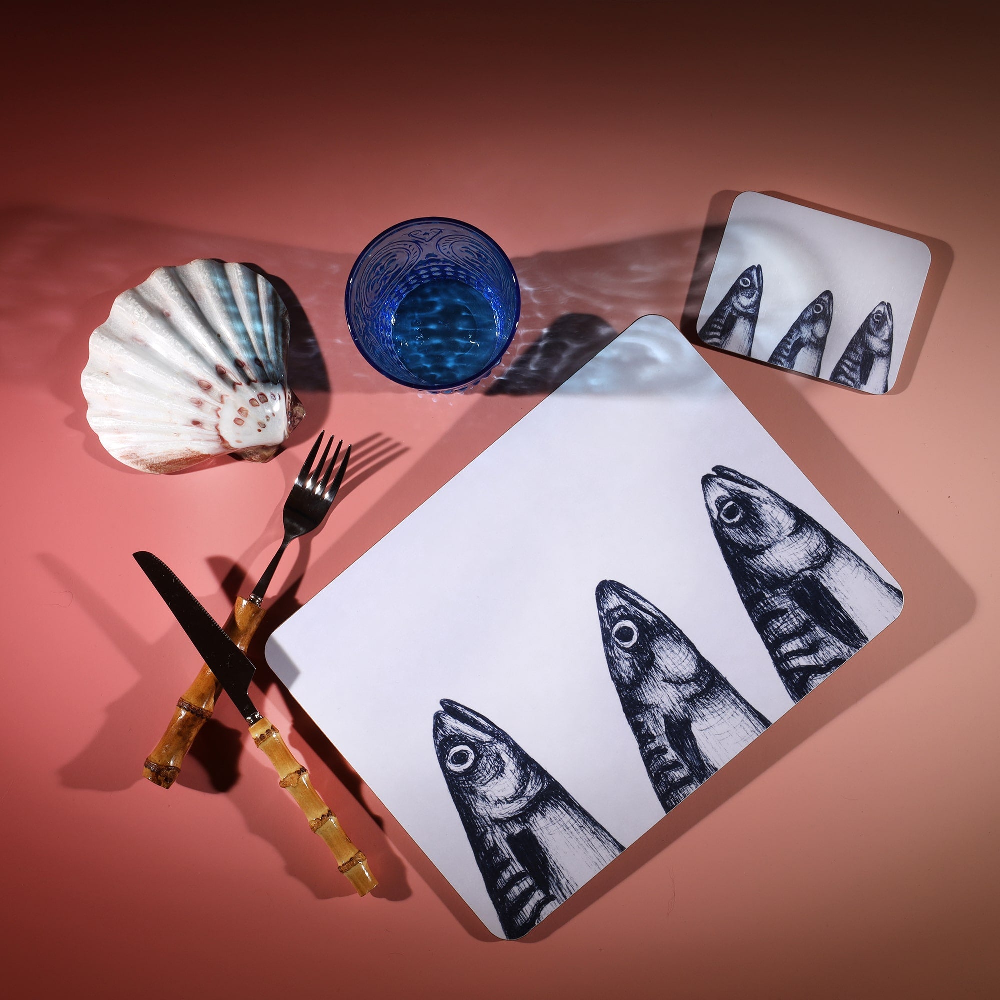 Mackerel Heads Design in Navy on a white Coaster with a matching Placemat.On the table is a bamboo cutlery set and a blue coloured glass