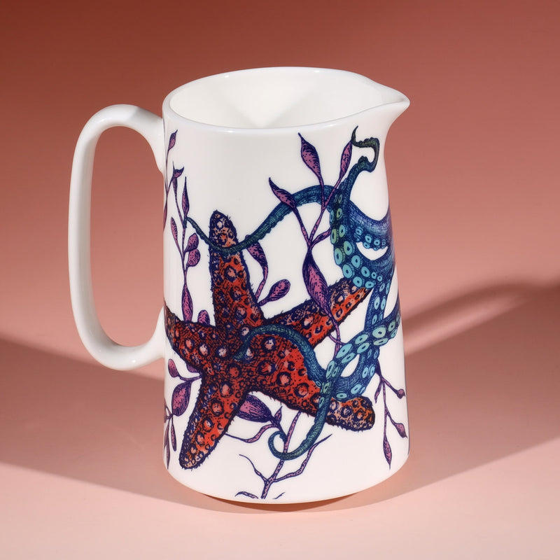 White jug with a brightly coloured blue, green orange and pink octopus tentacles starfish and seaweed design on a graduated coral coloured background. 