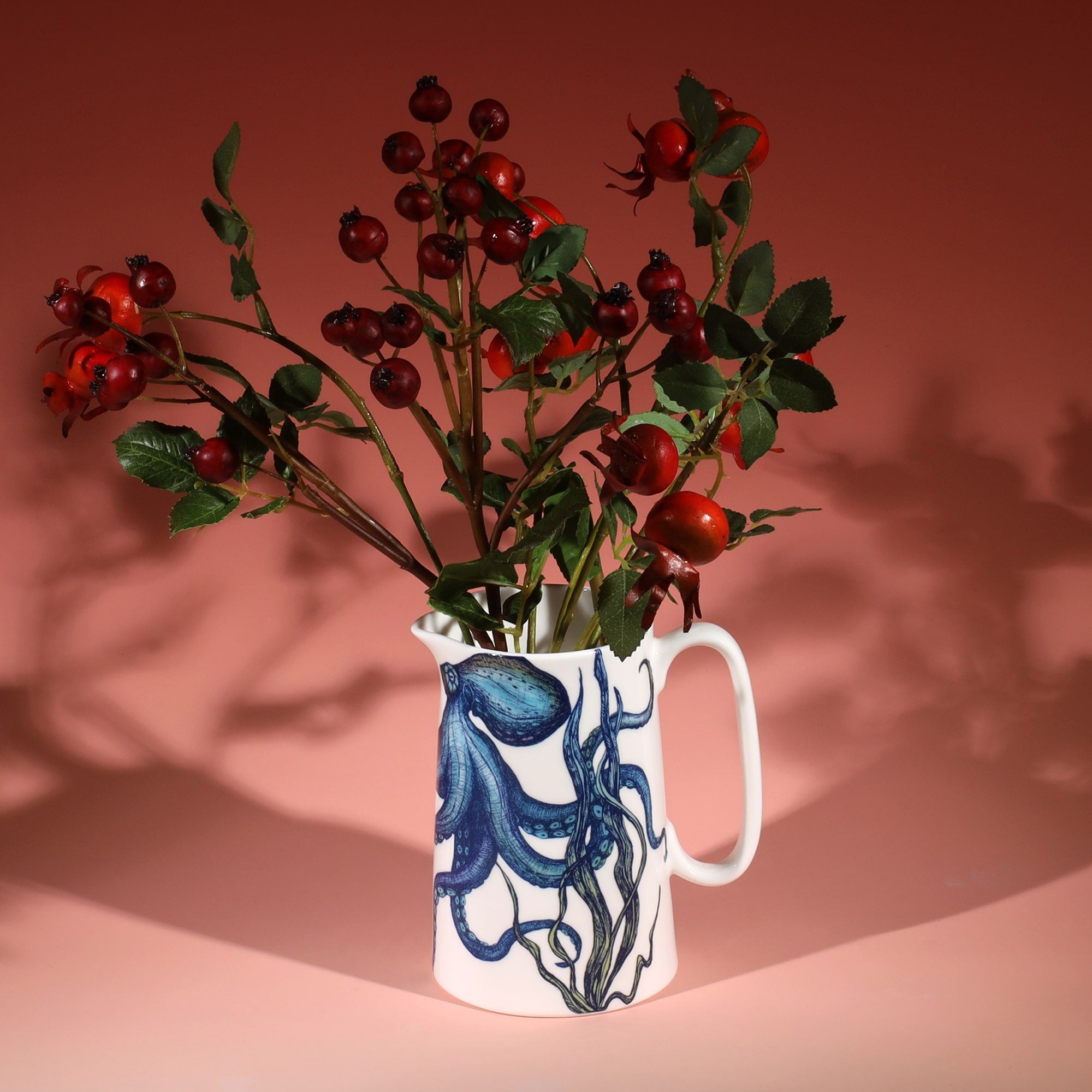 White jug with a brightly coloured blue and green octopus and seaweed design on a graduated coral coloured background with red rose hip branches in the jug.